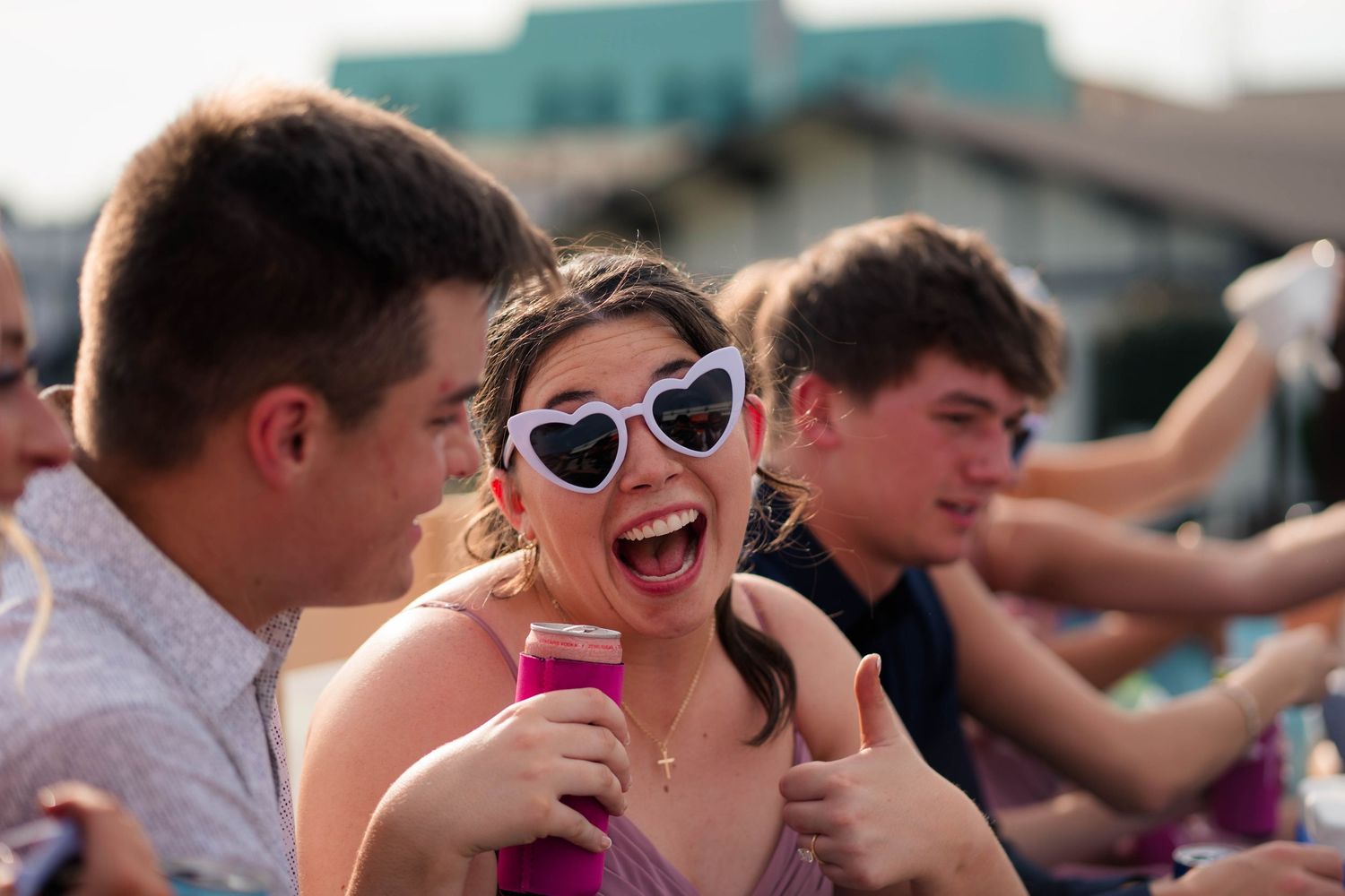 Excited young woman in heart-shaped sunglasses gives thumbs up while celebrating at an outdoor wedding reception.