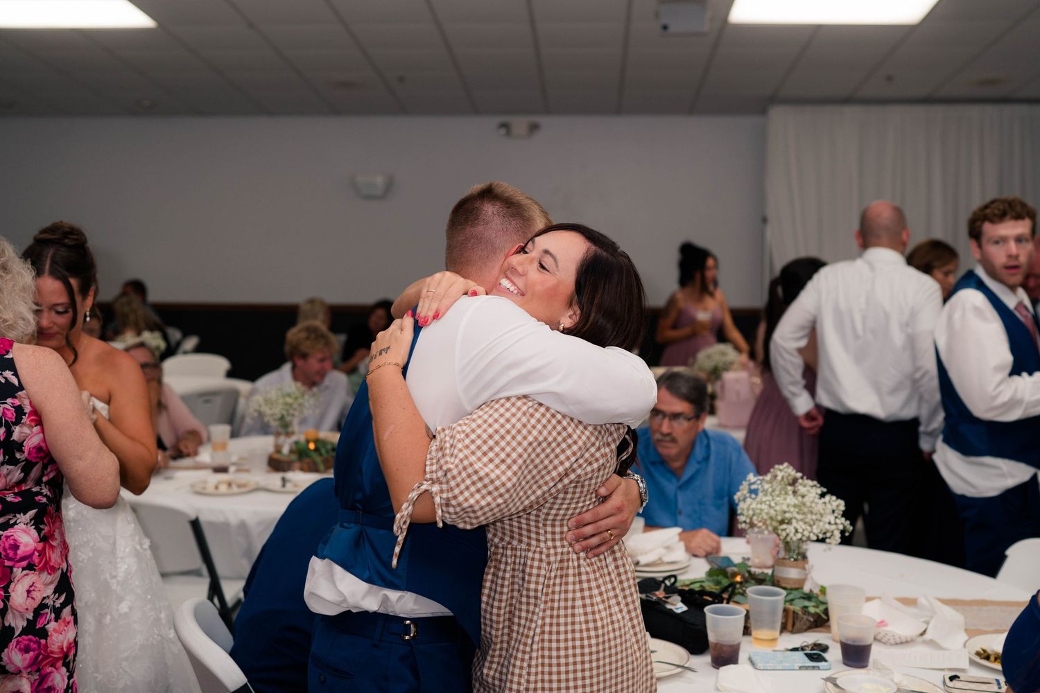 Two guests share a warm hug at a wedding reception, surrounded by elegantly decorated tables and seated attendees.