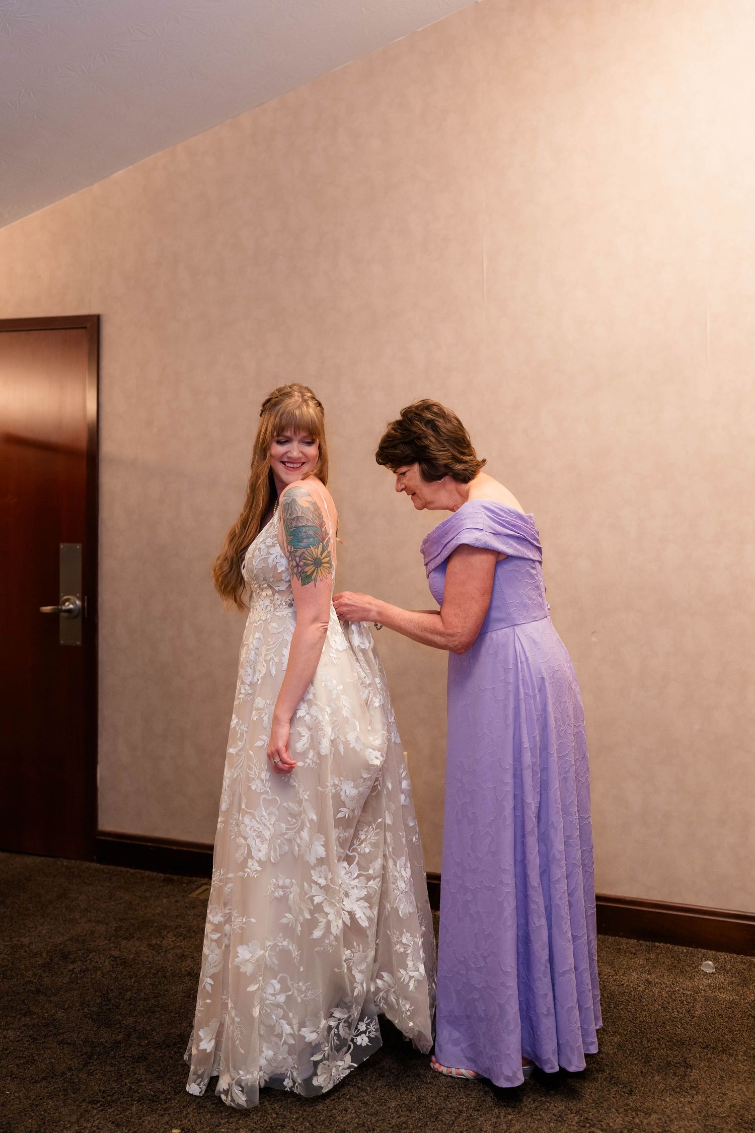 A bride in a floral lace gown smiles as a woman in lavender adjusts her wedding dress in a hotel room.