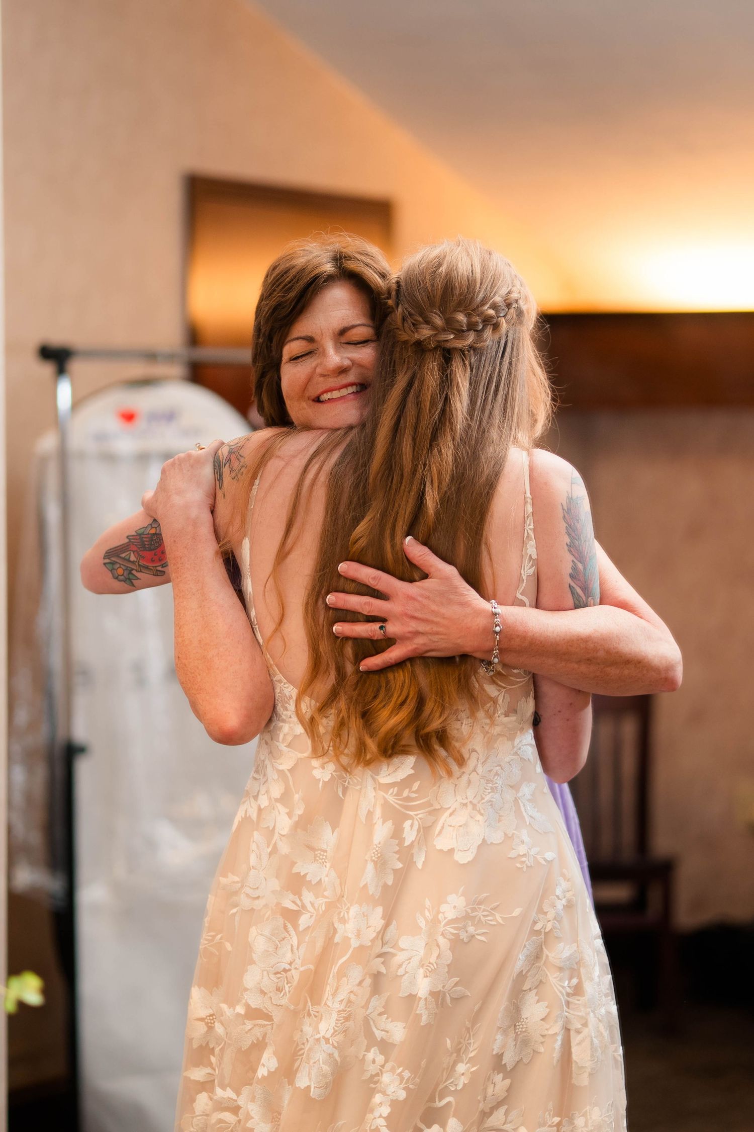 Two women share an emotional hug at a wedding, one in a floral lace bridal gown.