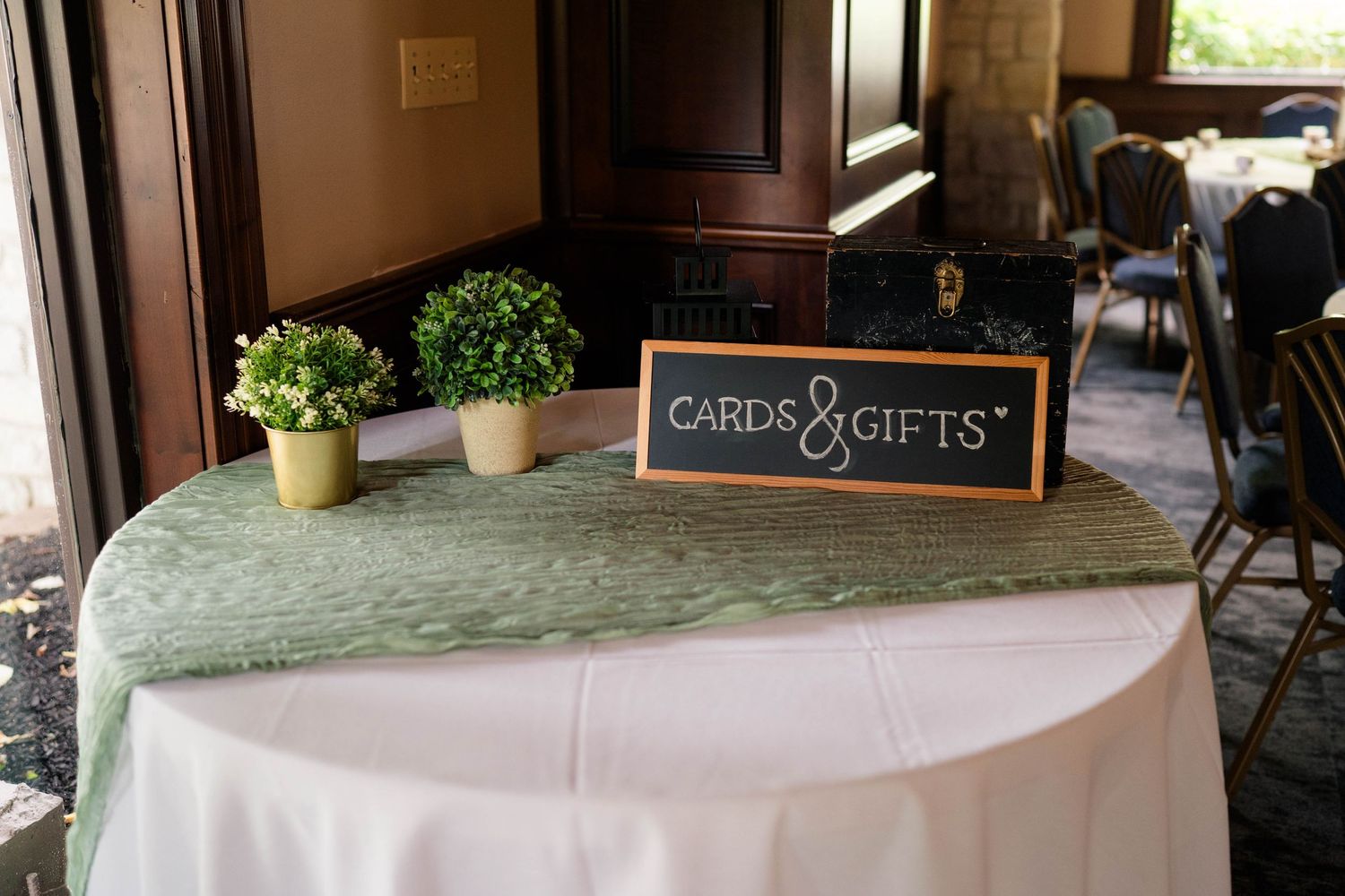Wedding cards and gifts table with green runner, sign, and decorative plants at reception.