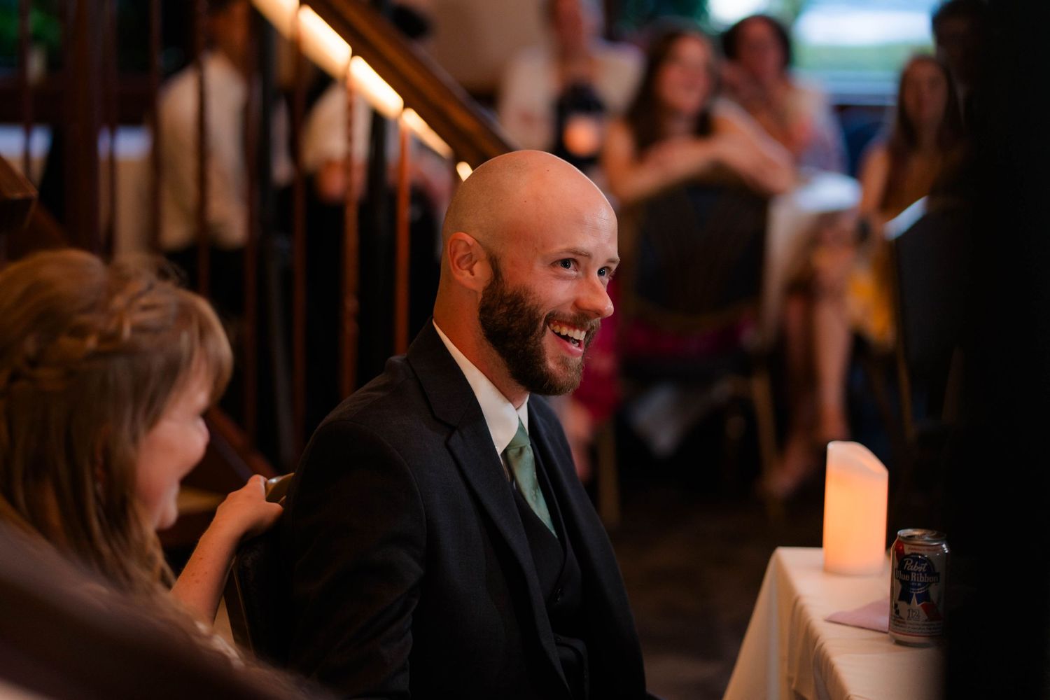 Bald groom smiling at wedding reception guests by candlelight.