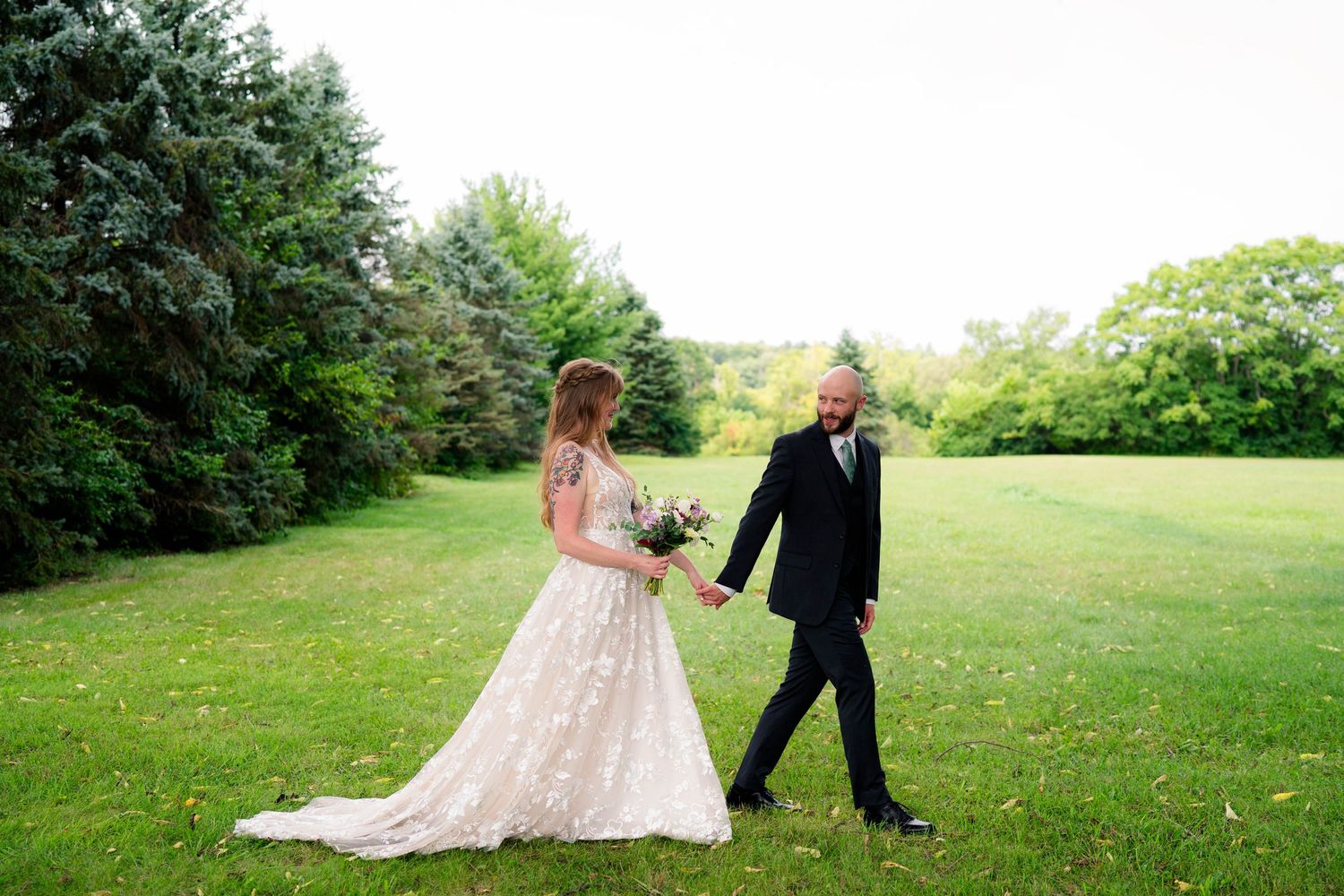 Bride in lace gown and groom holding hands walking across green lawn.