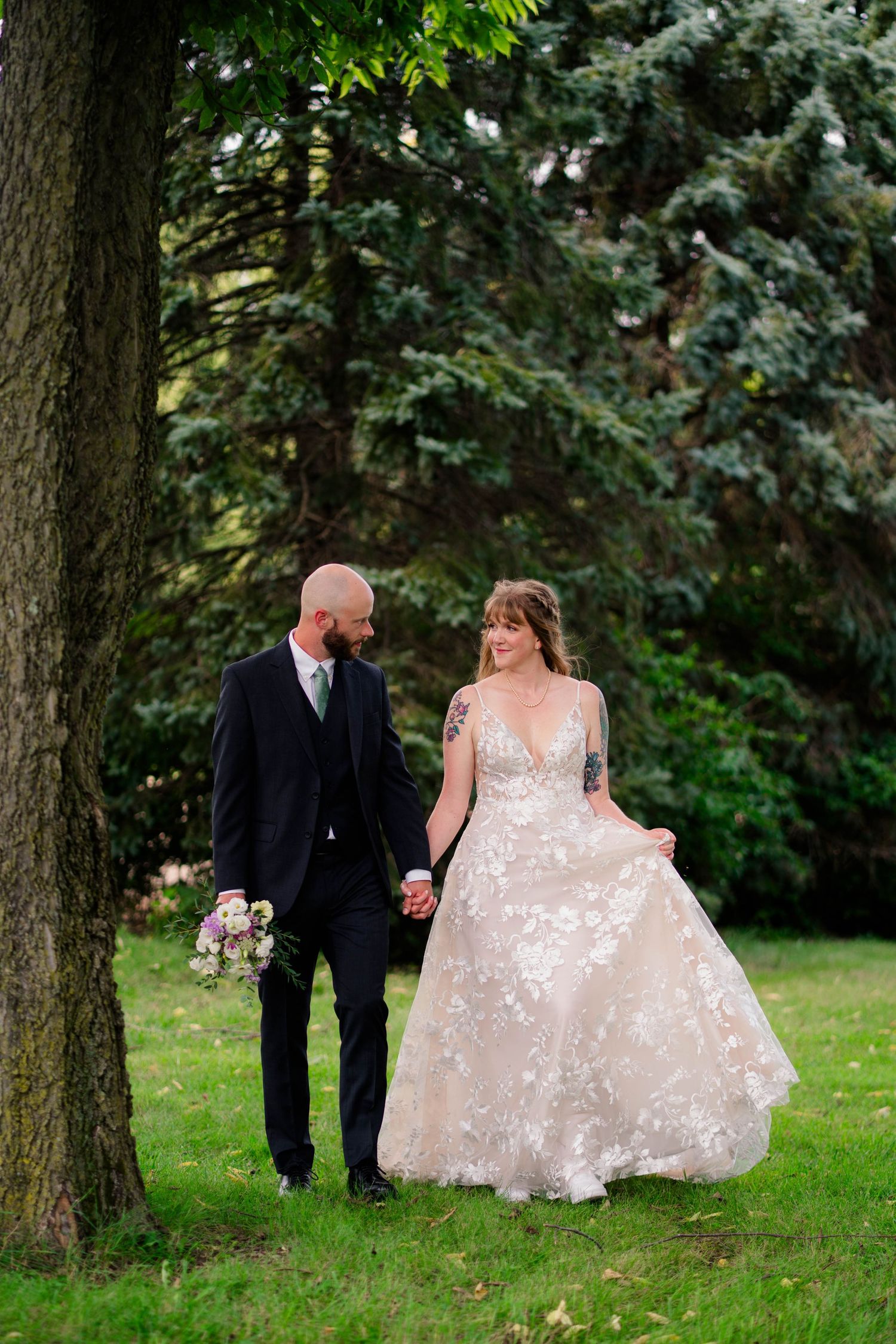 Bride and groom walk hand in hand through green park, bride in floral lace gown carrying bouquet.