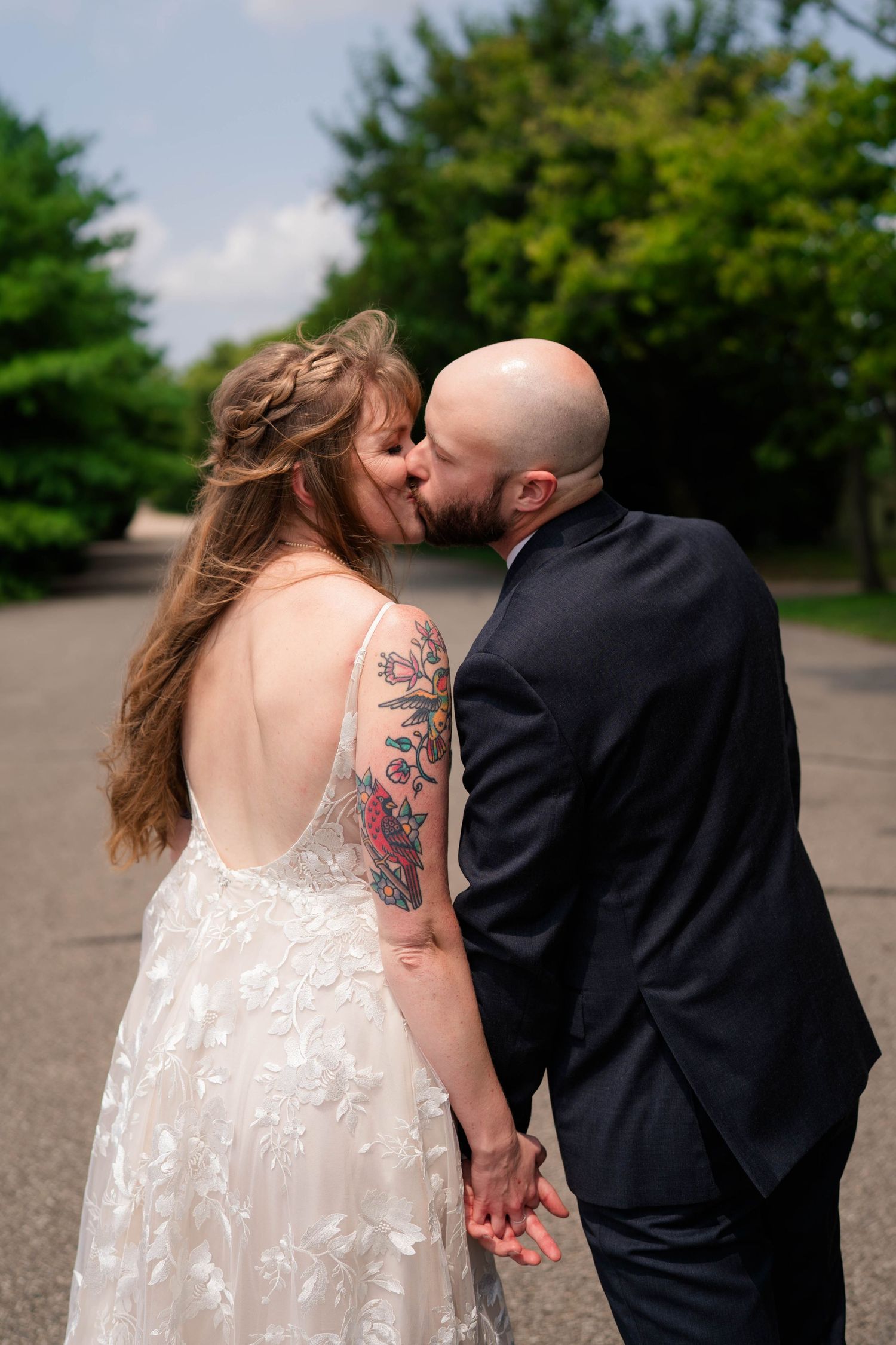 Tattooed bride and bald groom share a kiss outdoors on a sunny wedding day.