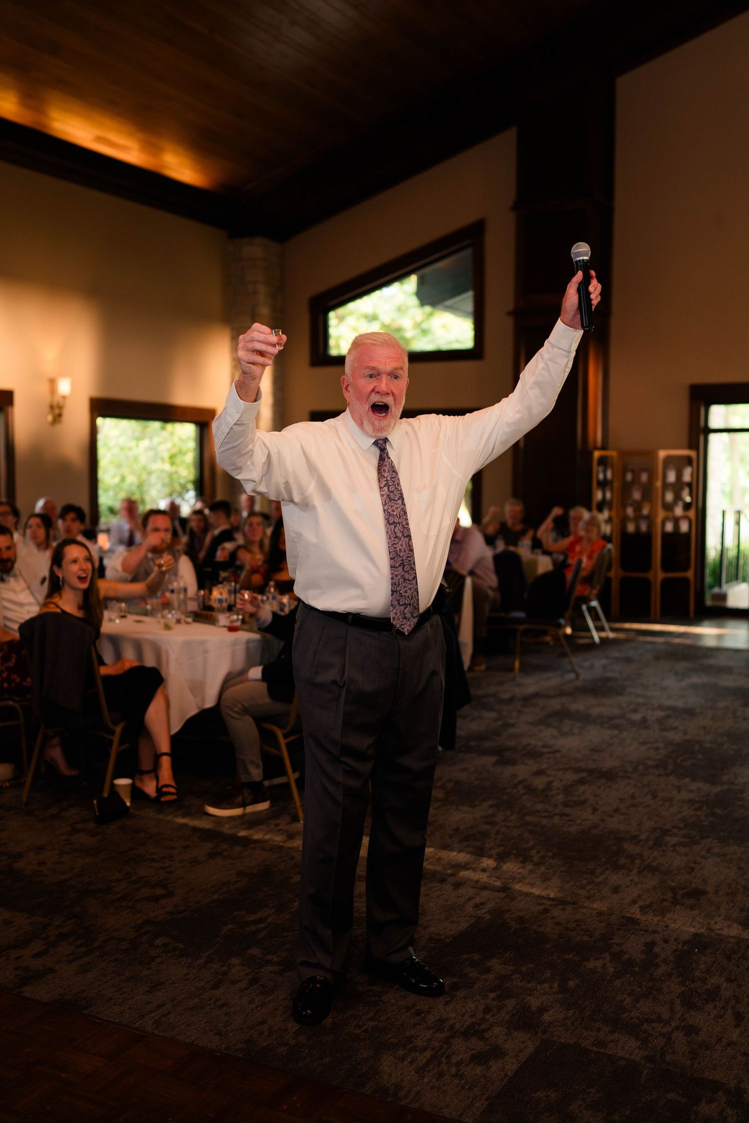 Elderly man celebrates enthusiastically with raised arms and microphone during wedding reception speech.