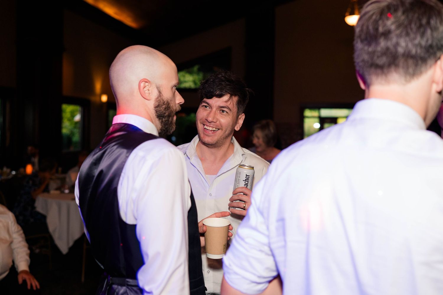Bald groom in vest smiles and chats with guests holding drinks at lively wedding reception.