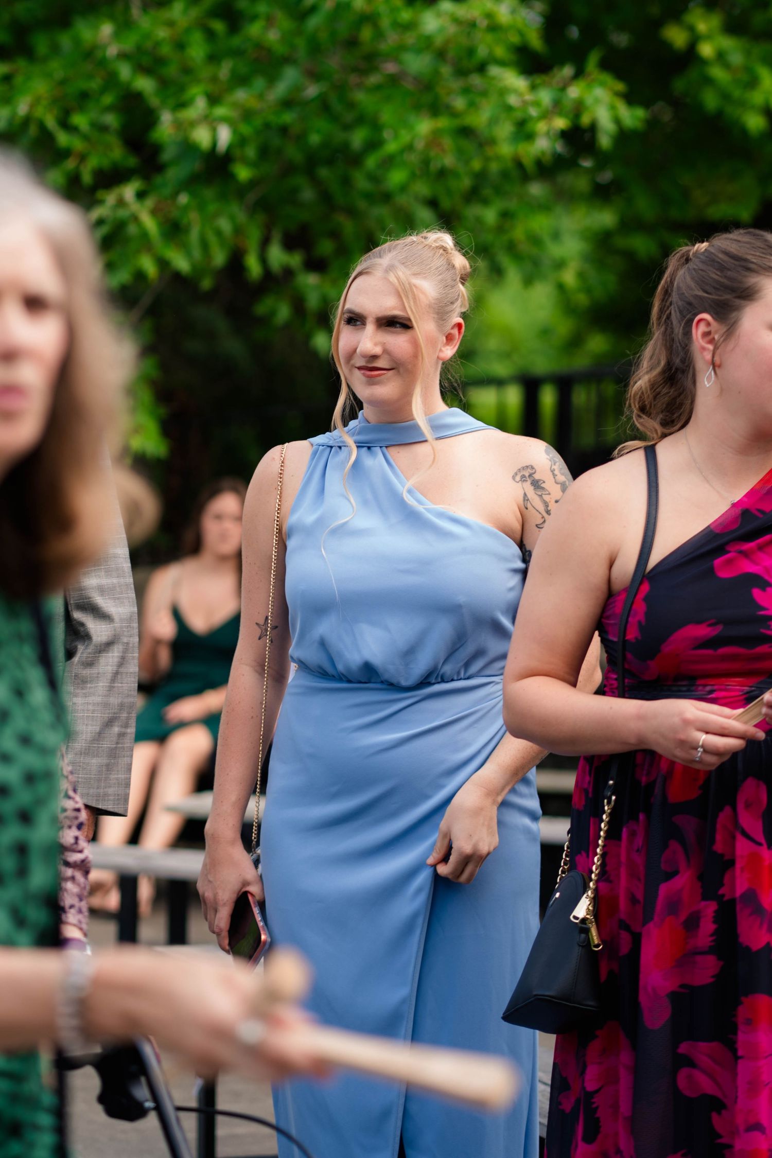 Elegant woman in blue halter gown with tattoos stands among wedding guests at outdoor ceremony.
