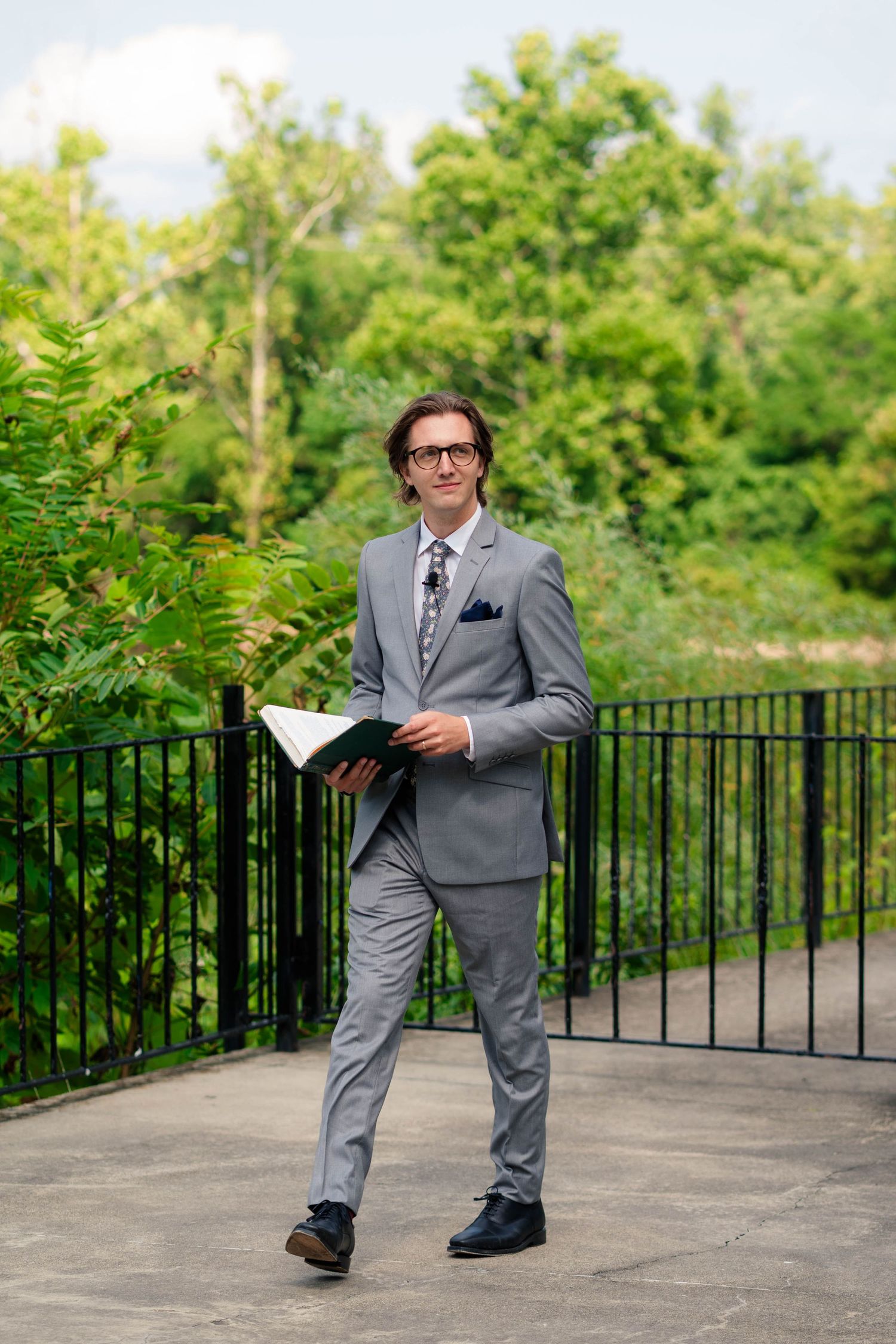 Elegant wedding officiant in a gray suit holds a ceremony book while standing outdoors near a black iron fence.