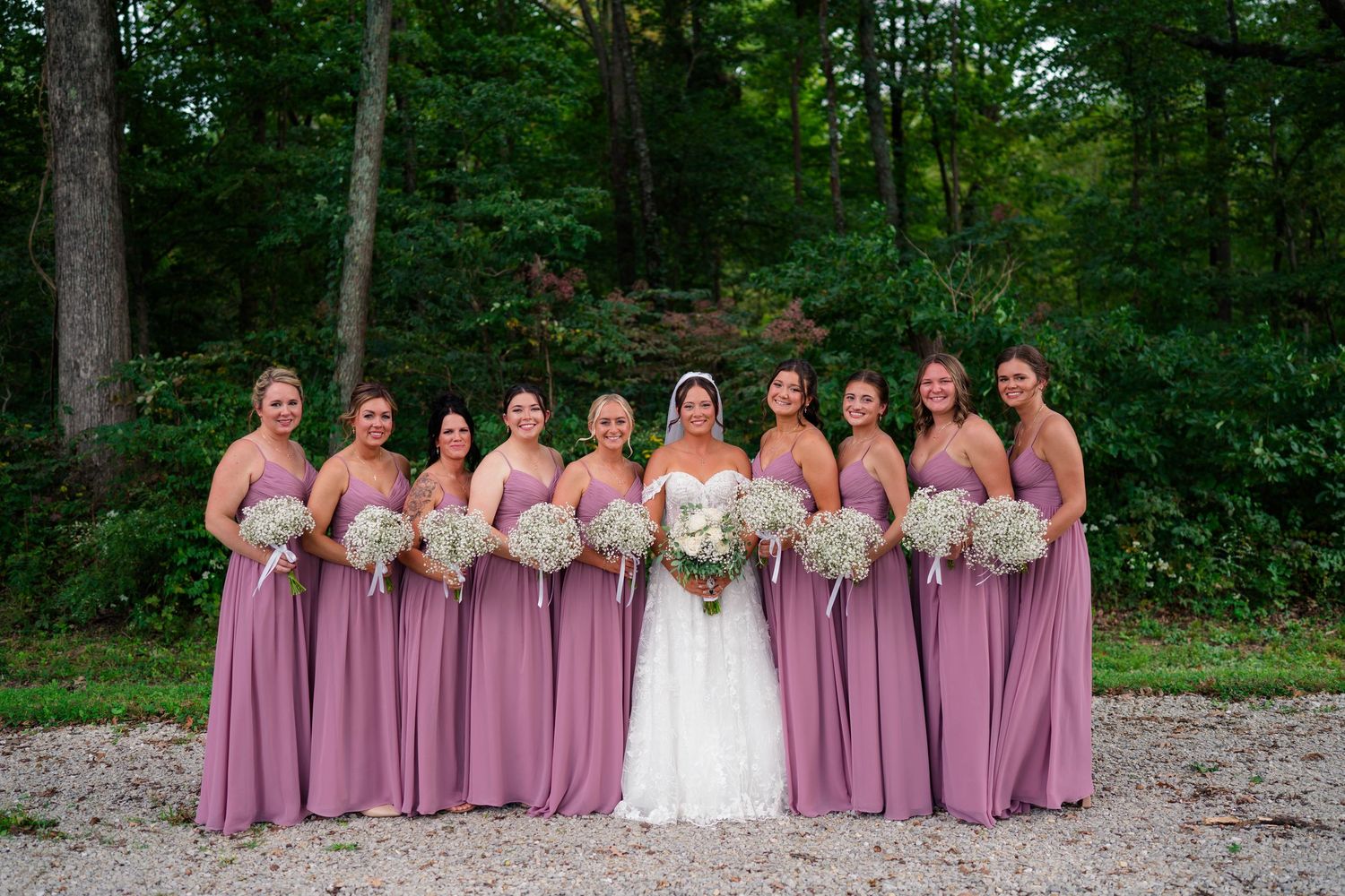 Bride poses with nine bridesmaids wearing matching mauve gowns holding white bouquets in a forest.
