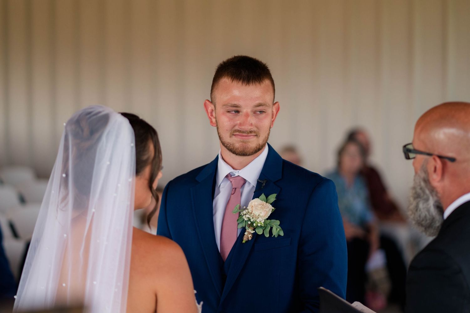 Emotional groom in navy suit watches bride approach during outdoor wedding ceremony.