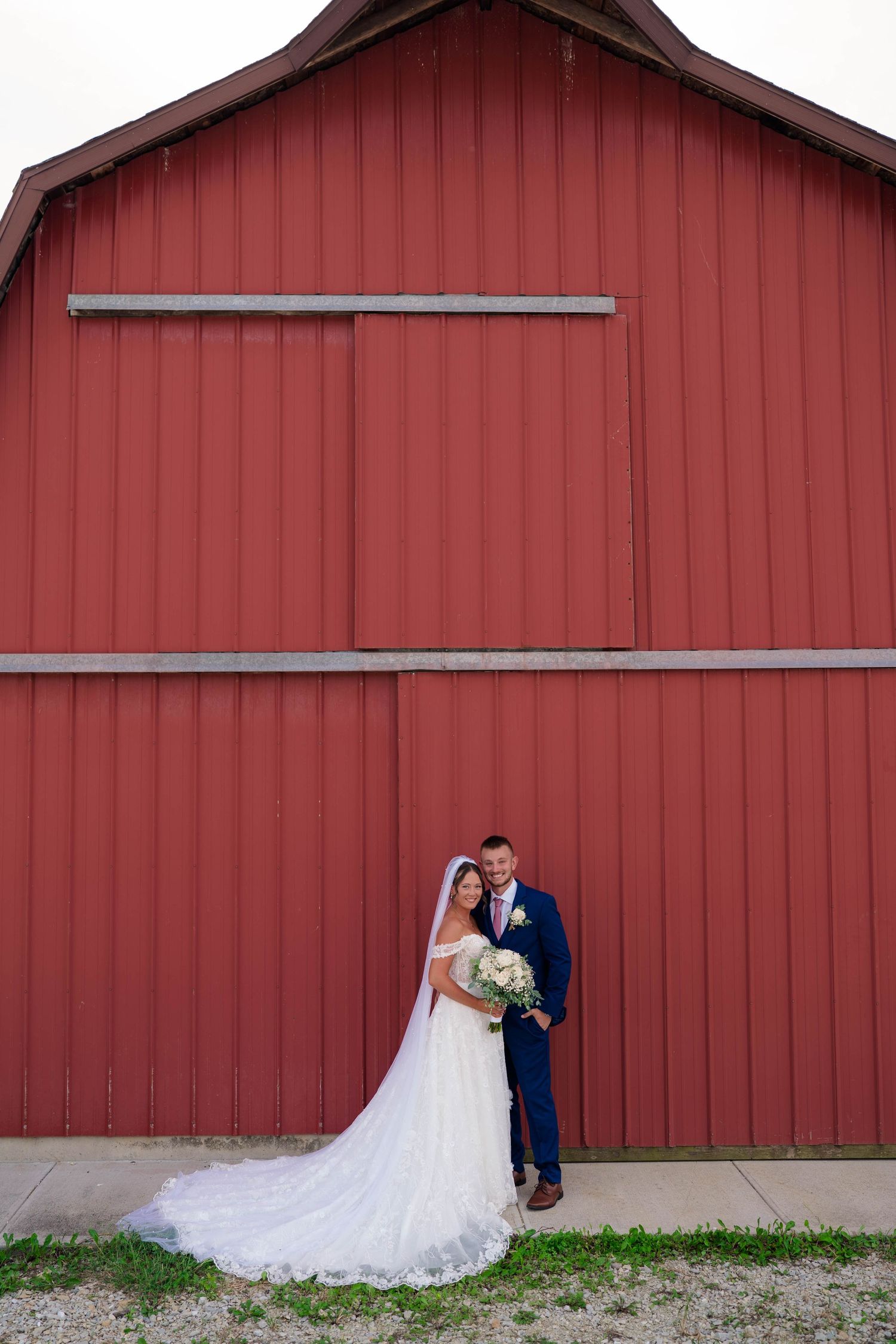 Bride and groom pose together in front of a large red barn, bride wearing a flowing white gown with veil.