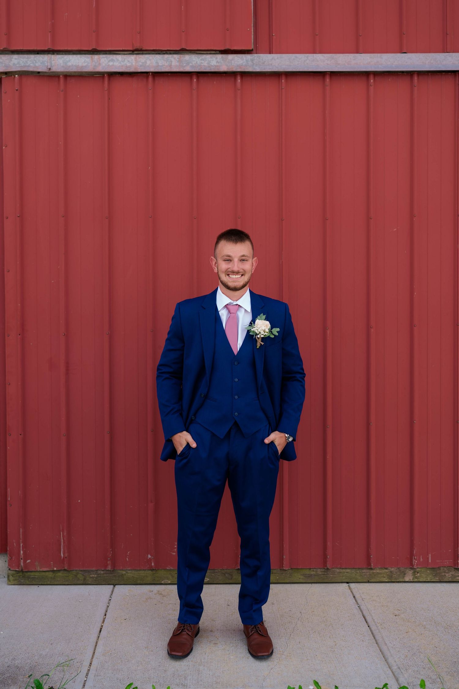 Smiling groom in navy blue three-piece suit with pink tie and boutonniere by red barn.