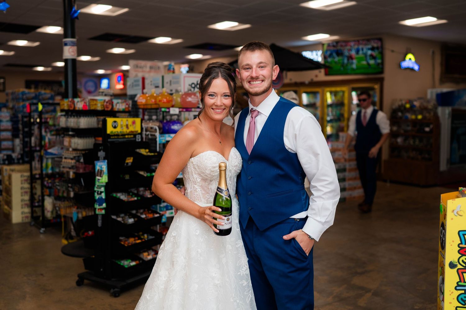 Smiling bride and groom pose with champagne bottle inside convenience store on wedding day.