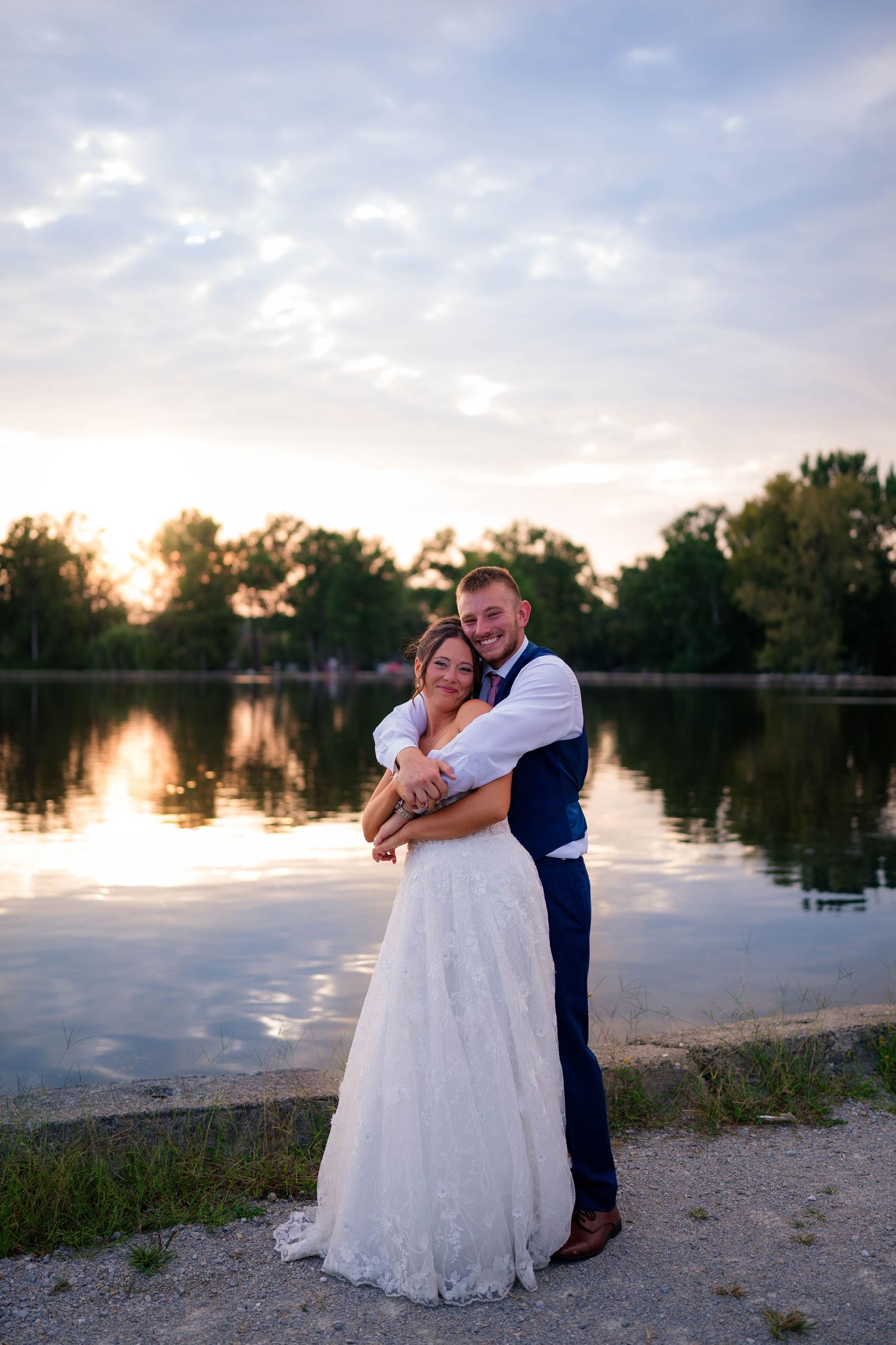 Newlywed couple embracing by a serene lake at sunset, bride in white gown and groom in navy suit.