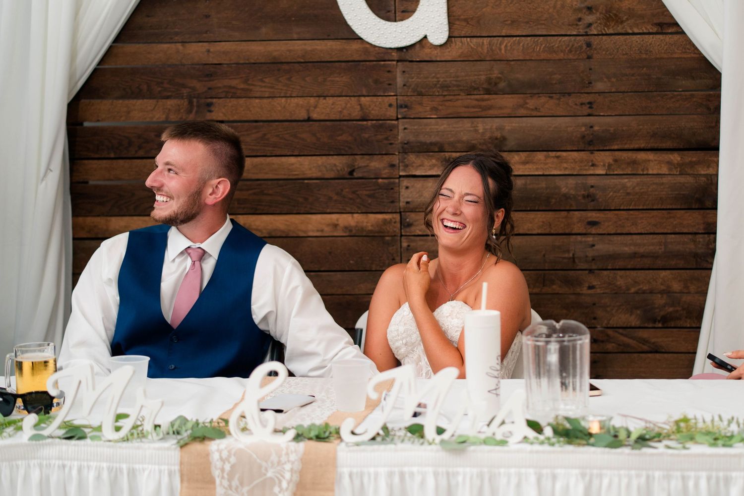 Bride and groom laugh joyfully at sweetheart table during wedding reception with wooden G monogram backdrop.