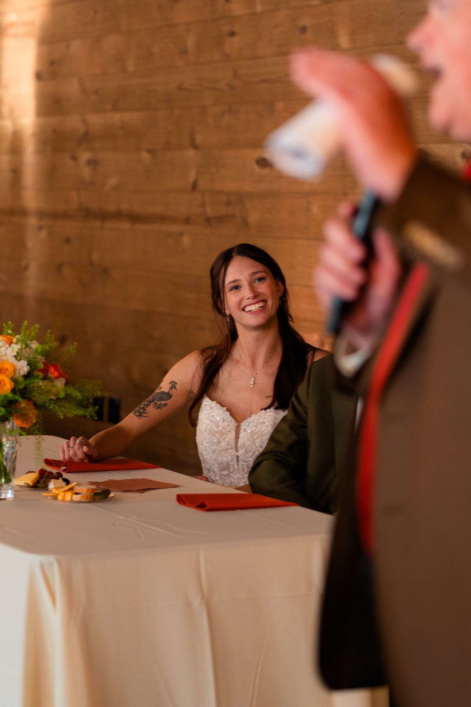 Smiling bride in lace gown with tattoos sits at sweetheart table during wedding reception toast.