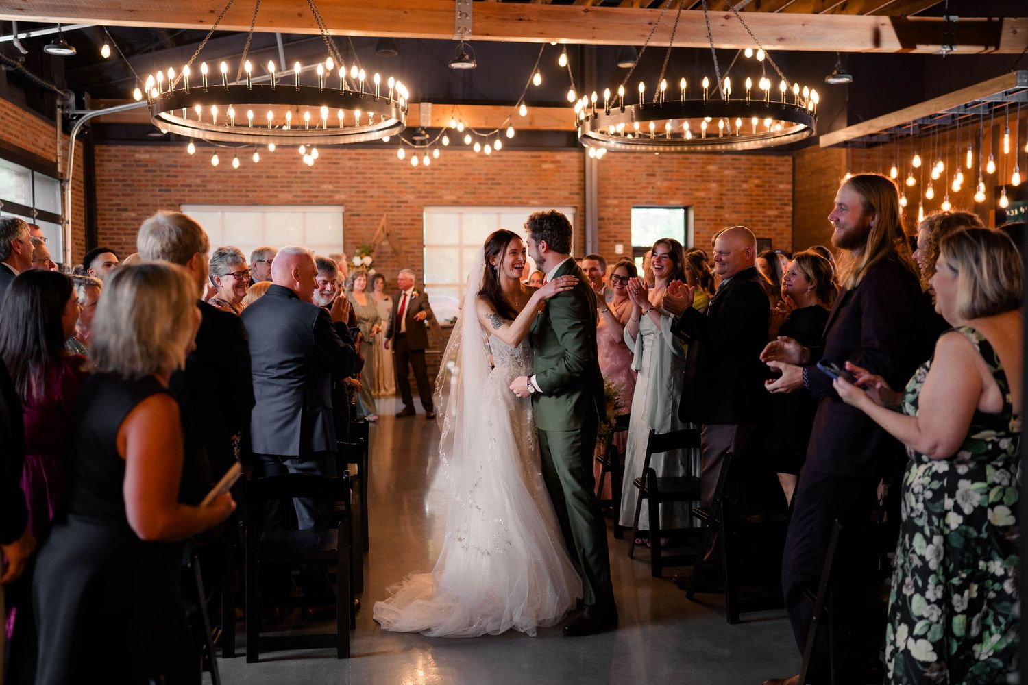 Newlyweds share first kiss as married couple in rustic industrial wedding venue with chandeliers.