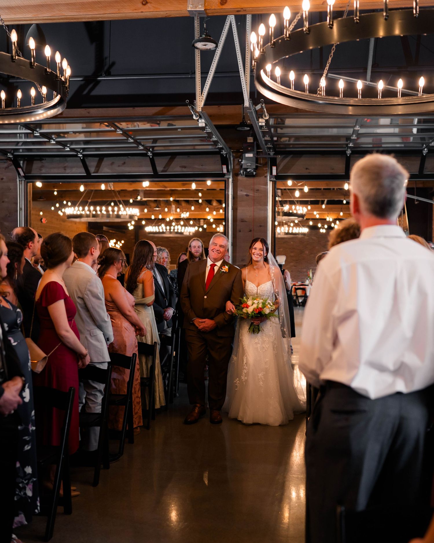Bride walks down aisle with escort at industrial wedding venue with Edison bulb chandeliers.