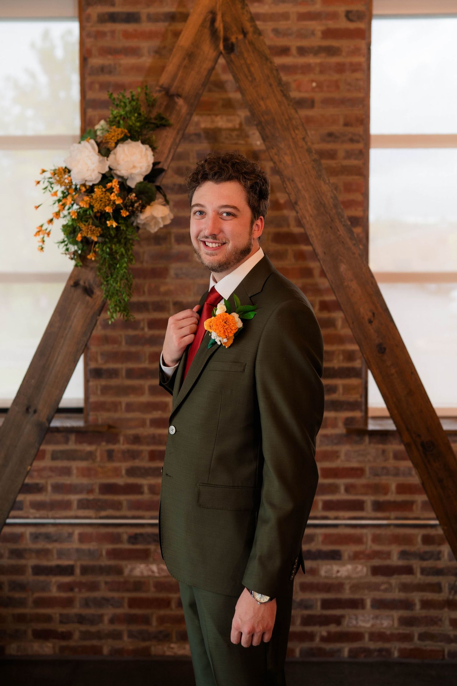 Smiling groom in olive suit with orange boutonniere posing before wooden arch backdrop.