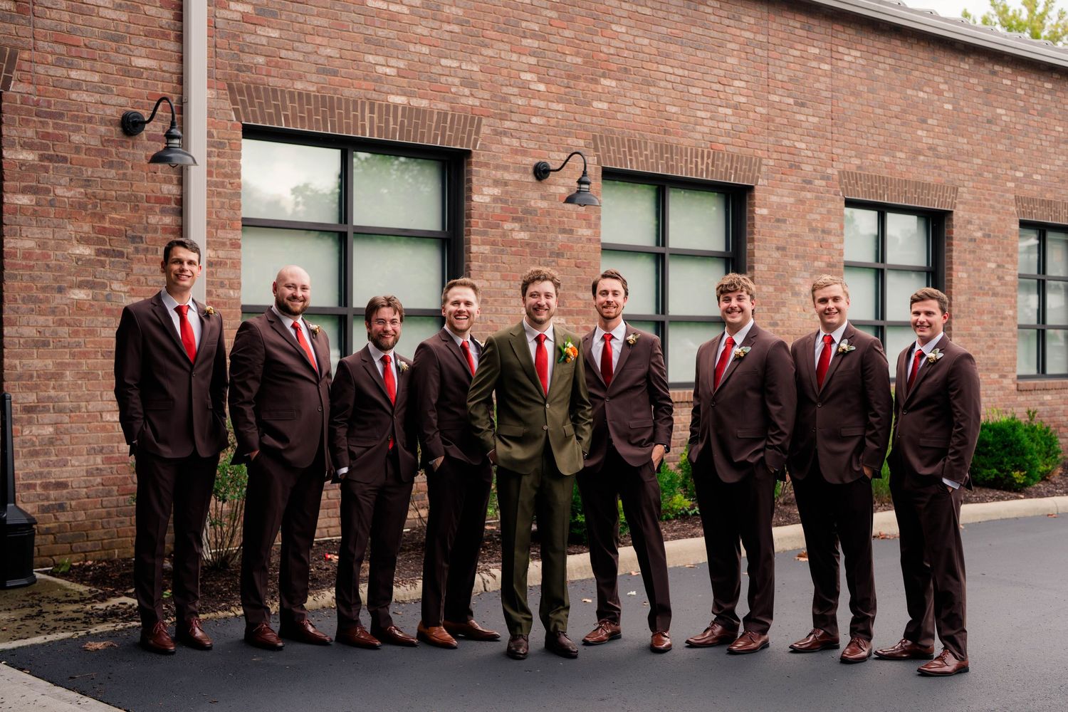 Group of groomsmen in dark suits and red ties posing outside brick wedding venue.