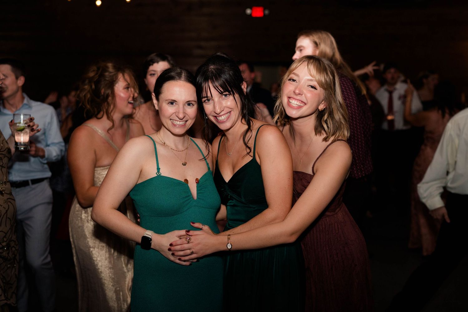 Three women in green and burgundy dresses smile together at wedding reception dance floor.