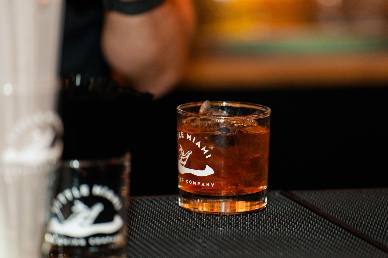 A whiskey cocktail with ice in a branded rocks glass on a dark bar surface.