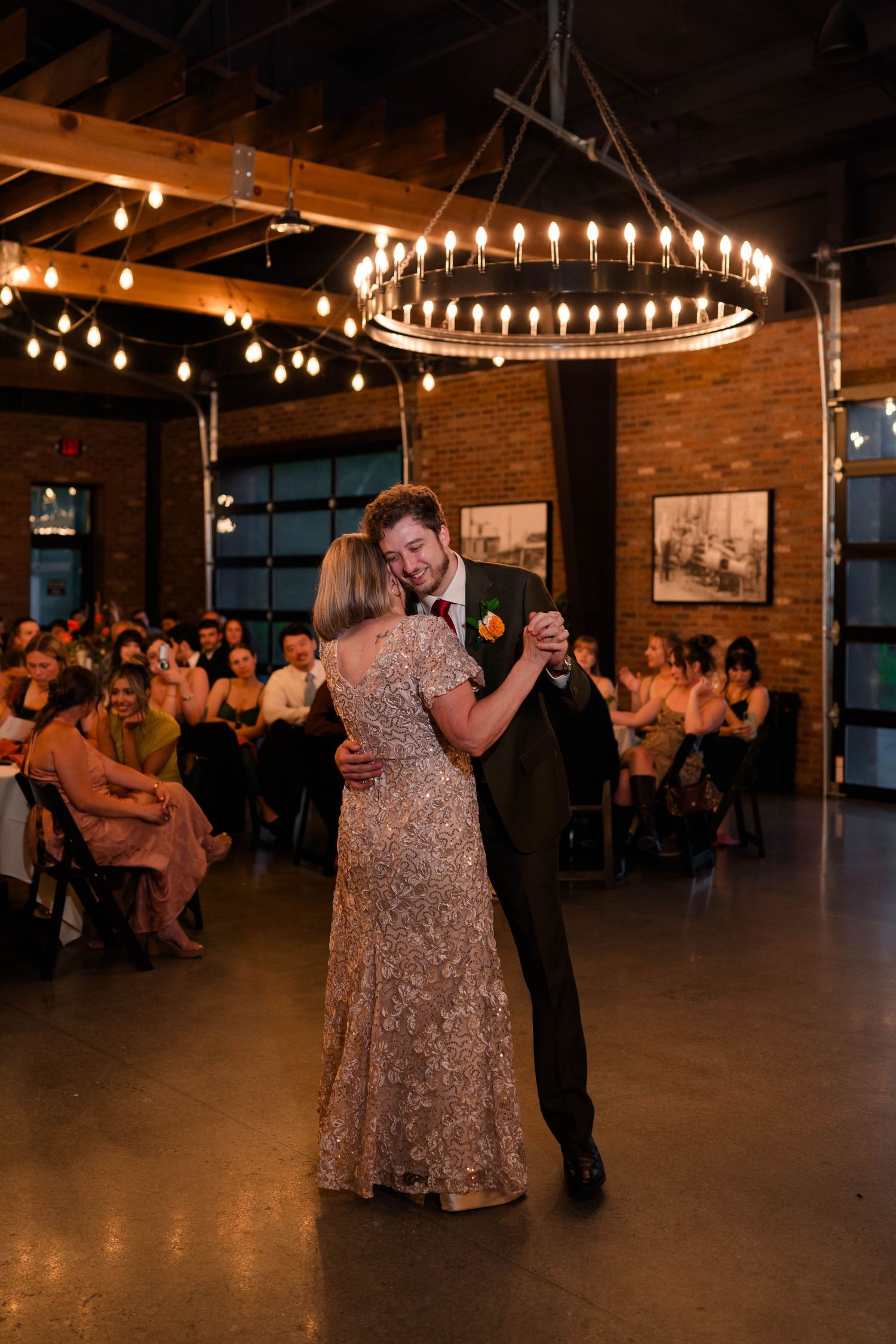 Groom dances with mother under elegant circular chandelier at warmly lit rustic brick wedding reception venue.