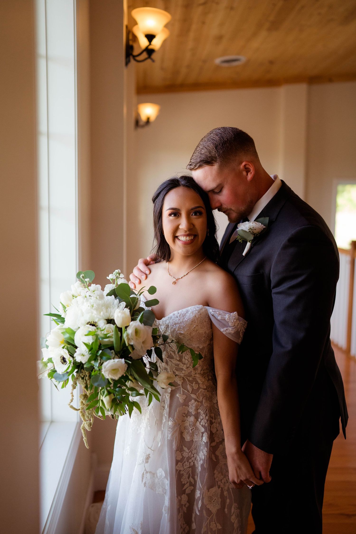 Bride smiles holding a white floral bouquet while groom embraces her tenderly indoors.
