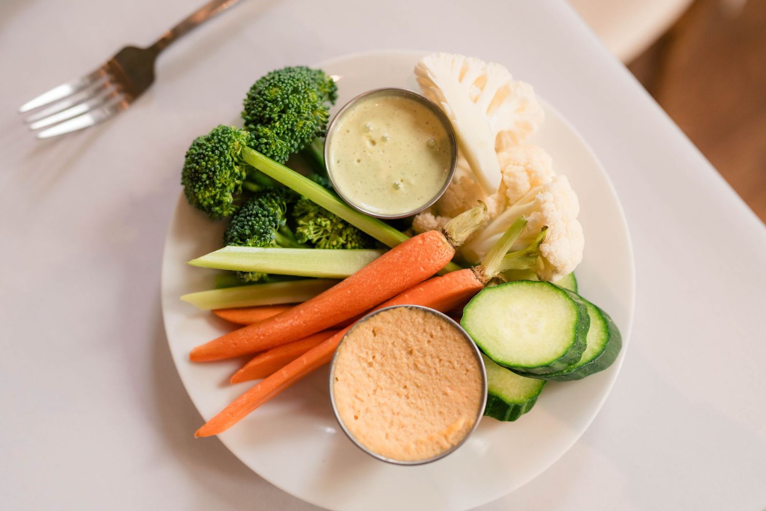 Fresh vegetable crudite platter with carrots, broccoli, cucumbers, and two dipping sauces on a white plate.