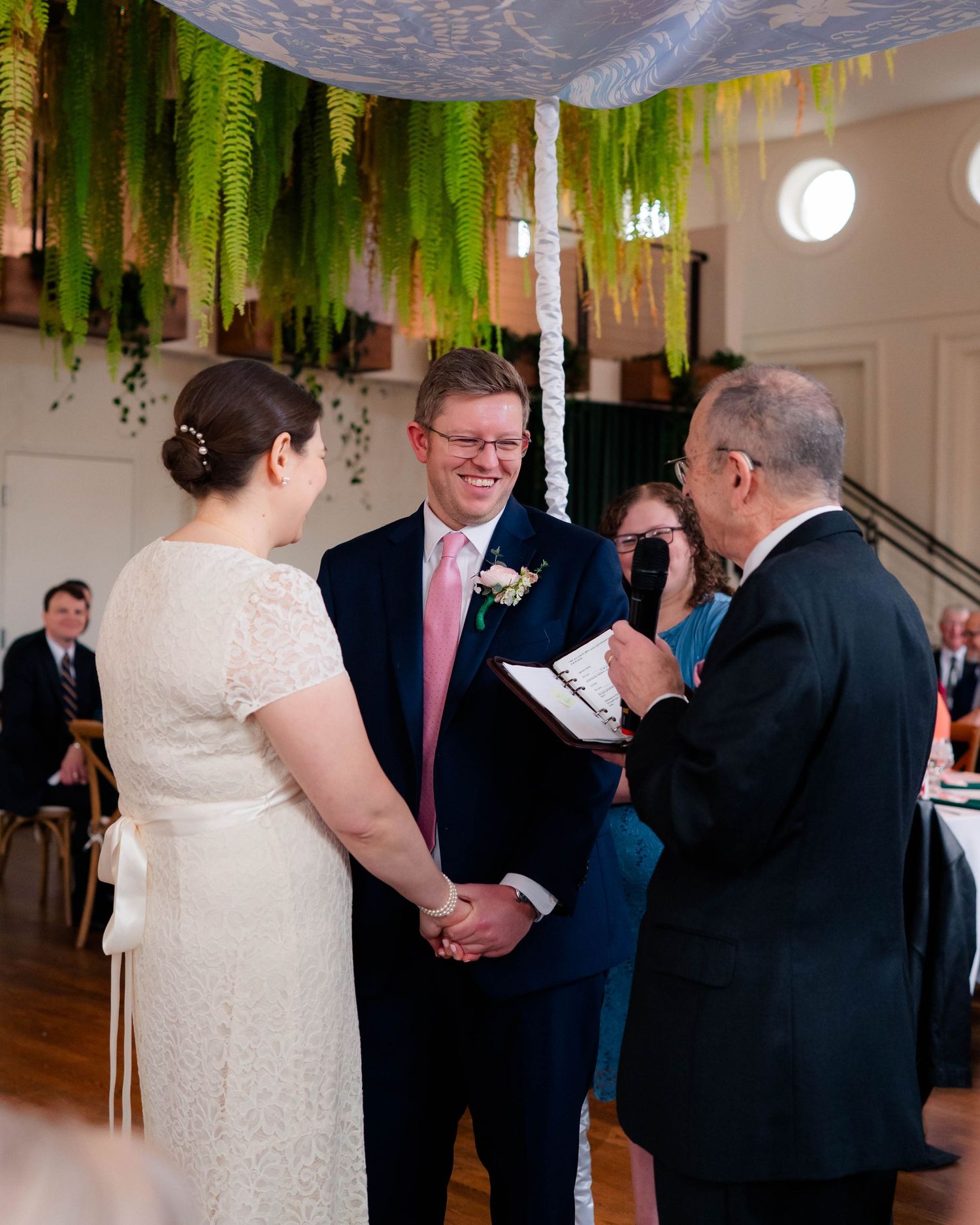 Smiling groom in navy suit holds bride's hands during Jewish wedding ceremony under chuppah with hanging ferns.