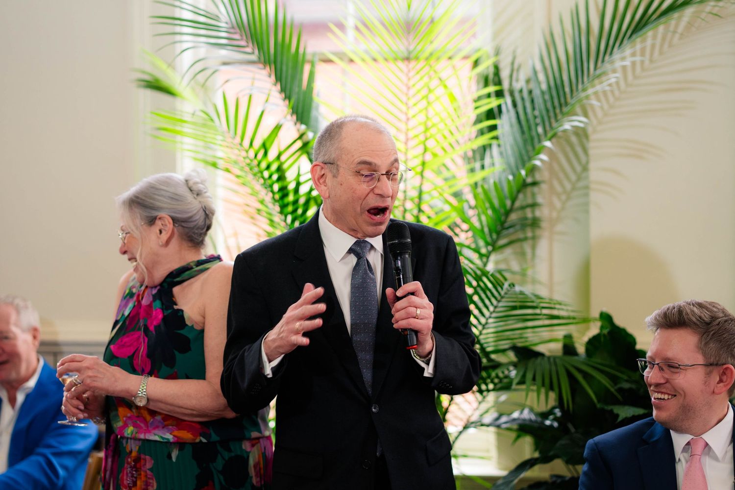 Man giving speech with microphone at elegant wedding reception with guests laughing in background.