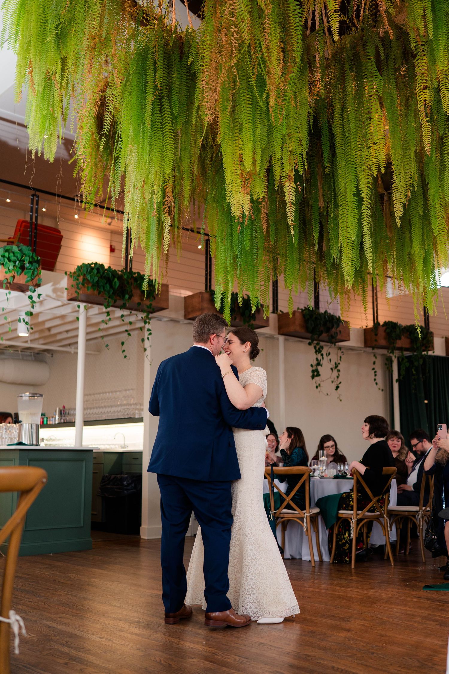 Newlyweds embracing under lush hanging ferns during first dance at botanical reception.