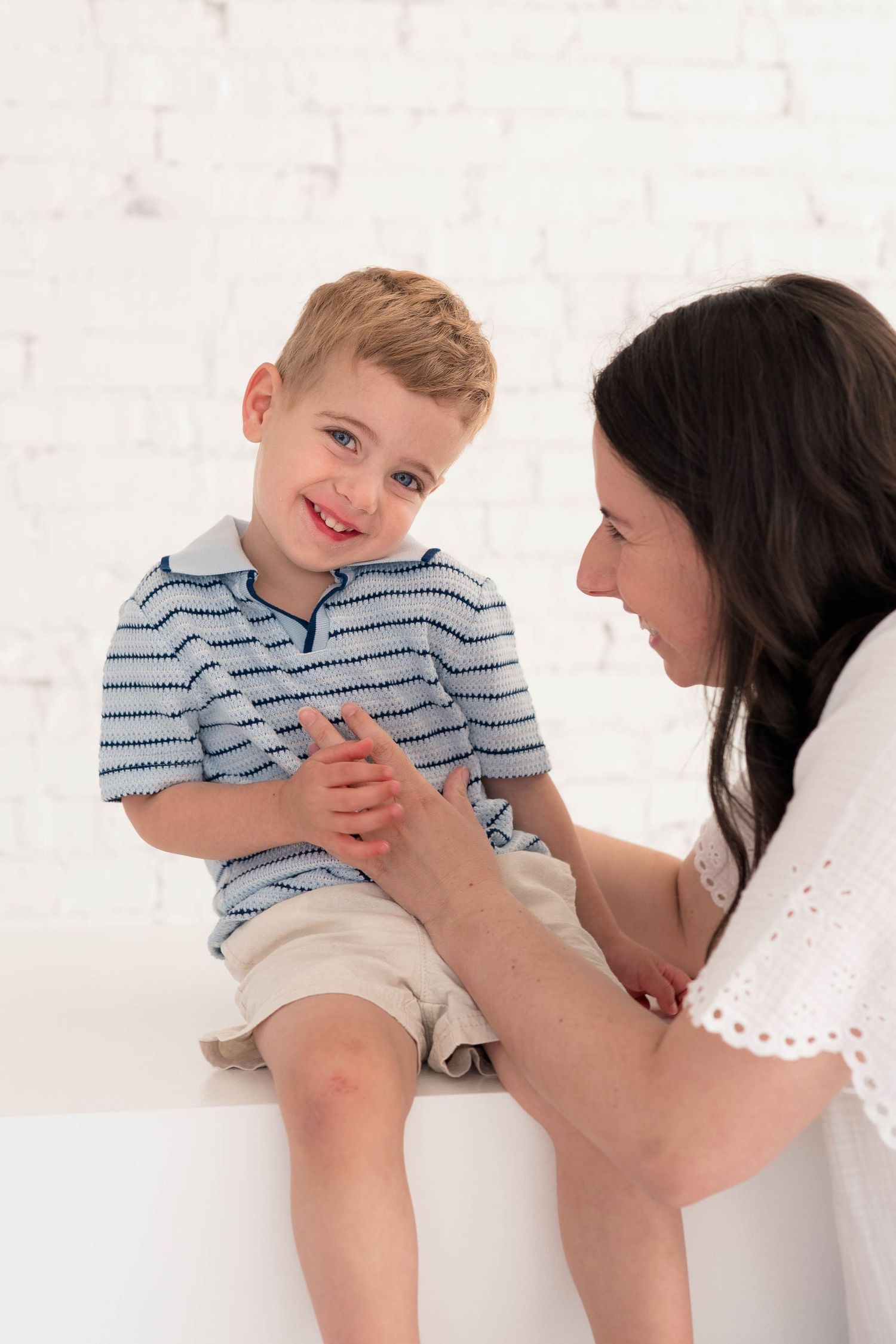 A smiling blonde toddler boy sits on a white surface while his dark-haired mother tenderly holds his hands and smiles at him.