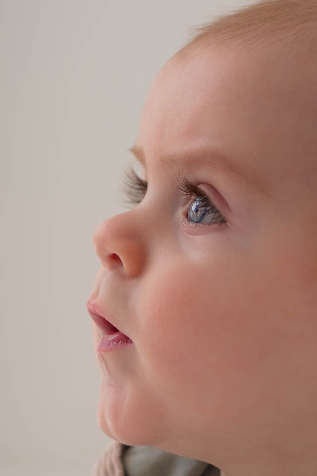 Close-up portrait of a curious baby with blue eyes and blonde hair looking upward against a light background.