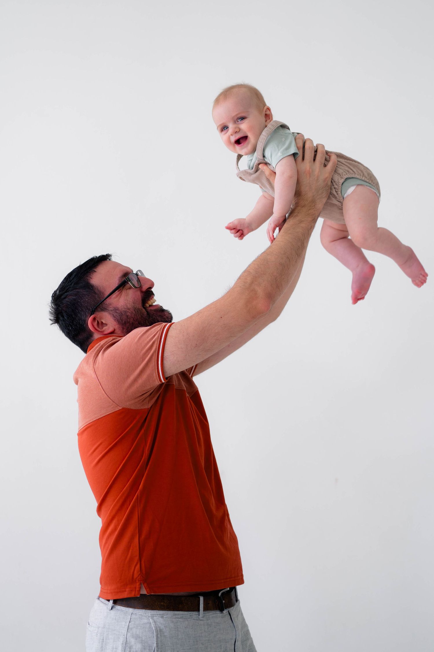 Father in orange polo shirt joyfully lifting smiling baby overhead against white background.