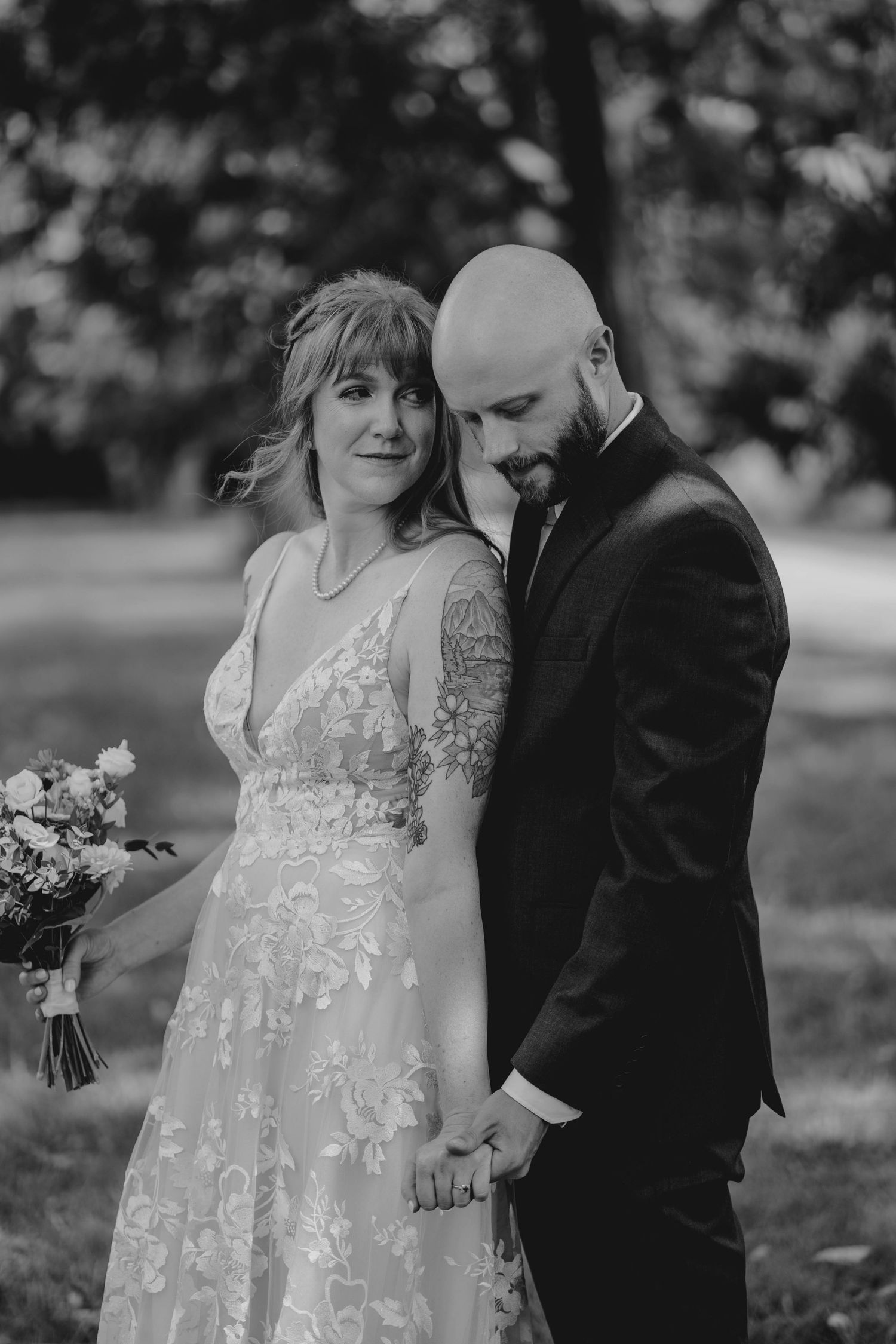 Black and white portrait of a bride and bald bearded groom holding hands tenderly in an outdoor setting.