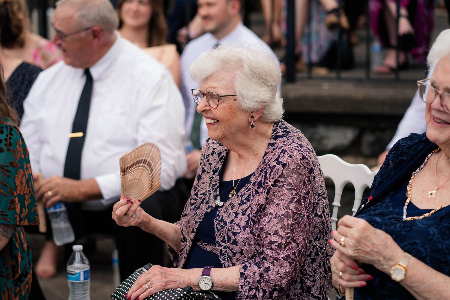 Elderly woman smiles and fans herself while seated among guests at an outdoor wedding reception.