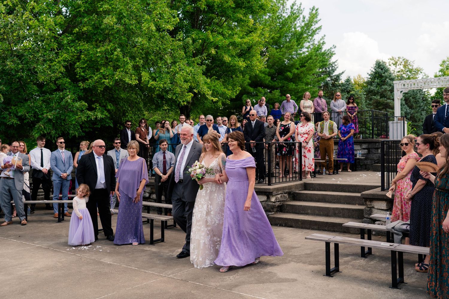 Bride walks down outdoor aisle with wedding party in lavender dresses during sunny ceremony.