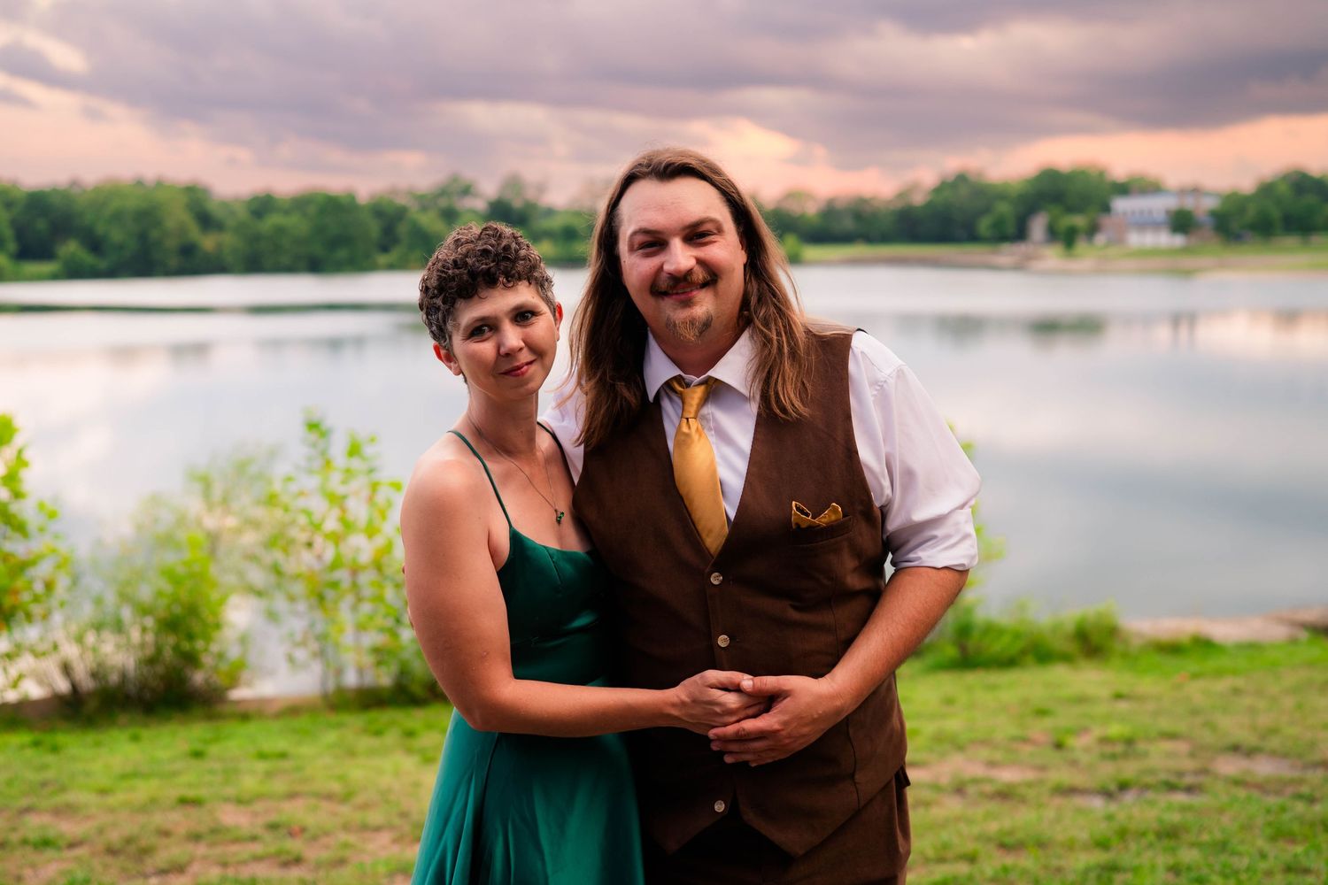 Smiling couple poses outdoors by a serene lake at sunset, man in brown vest and woman in emerald green dress.