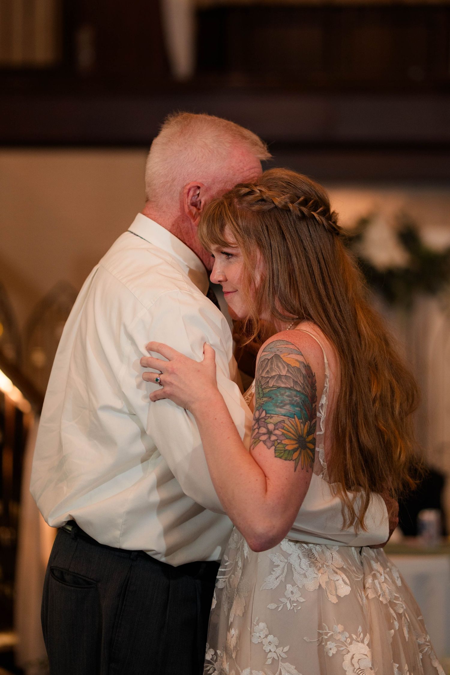 Bride with braided updo and floral tattoo shares an emotional embrace during father-daughter wedding dance.