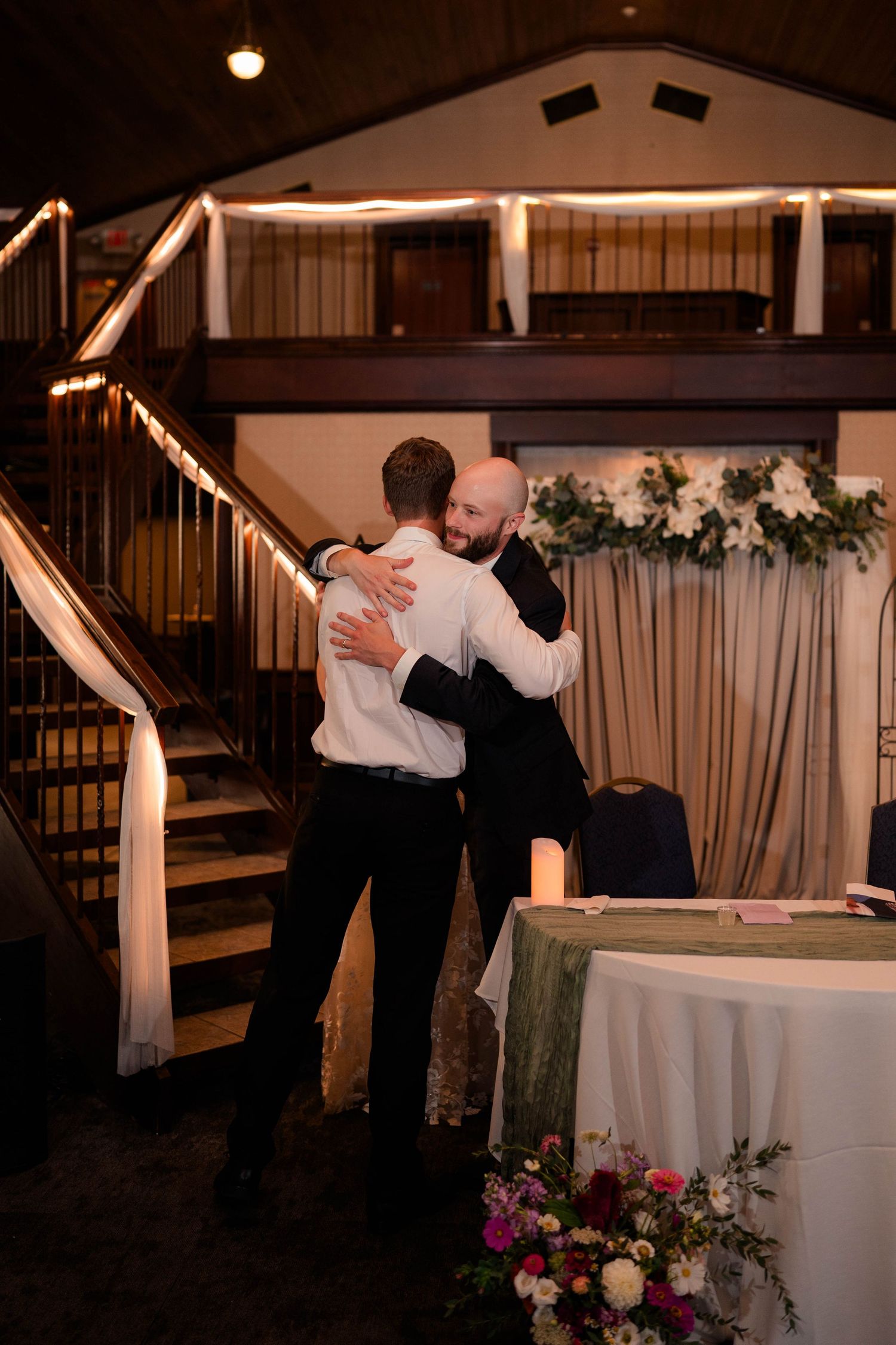 Bald groom embraces a guest in an emotional hug inside a warmly lit wedding reception hall.