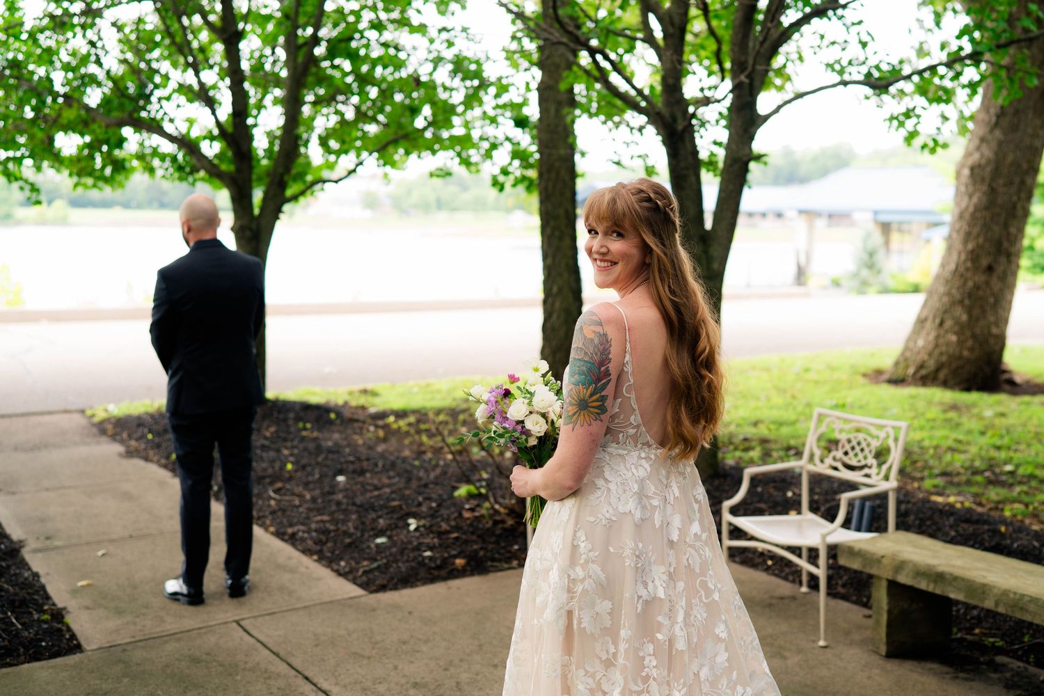 Bride in lace wedding gown with floral tattoo holds bouquet during outdoor first look moment.