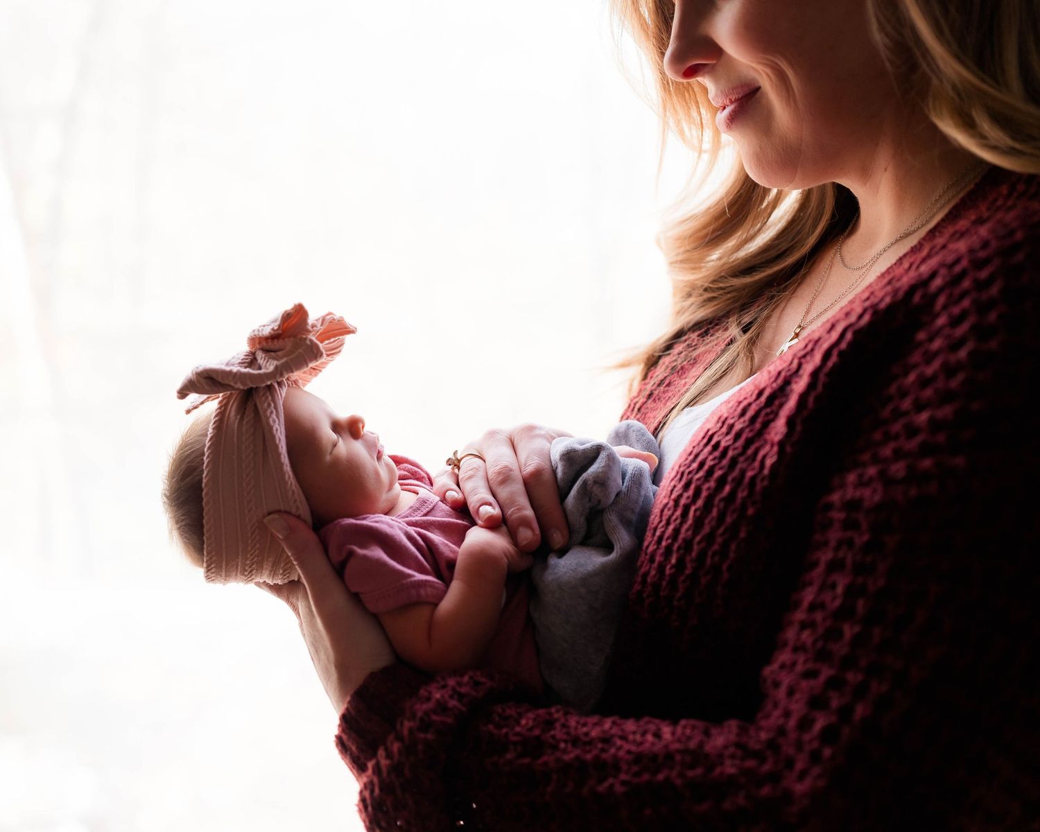 A smiling blonde mother cradles her sleeping newborn daughter in a pink outfit and bow headband in soft natural light.