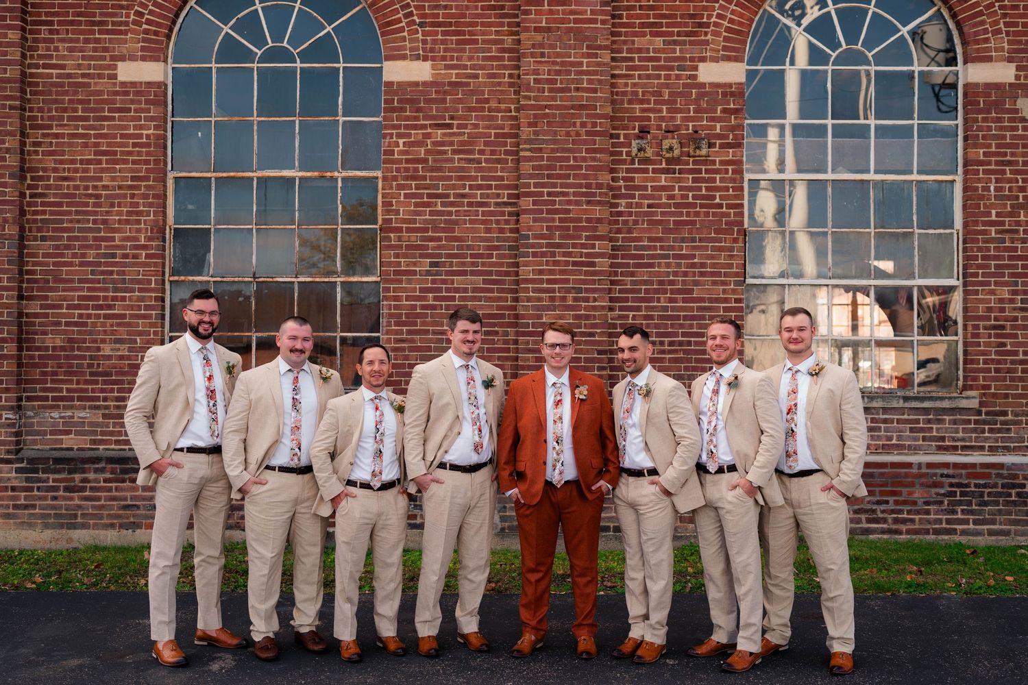 Groom in rust suit poses with groomsmen in tan suits before a brick building.
