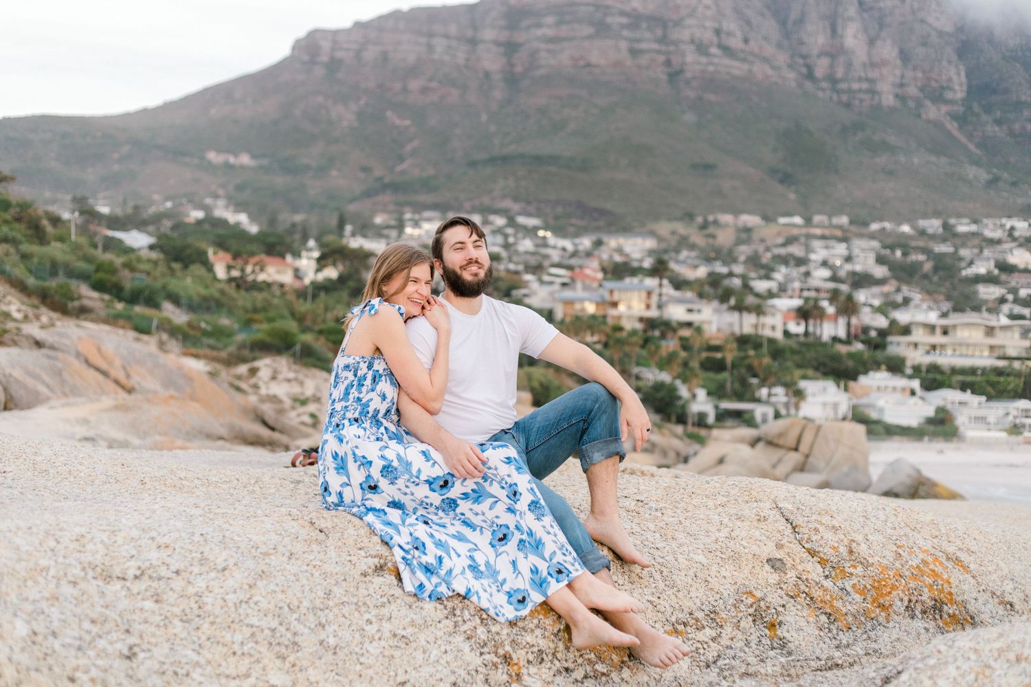 Couple sitting on rocks at Clifton Beach, Cape Town, with mountain and coastal town in background.