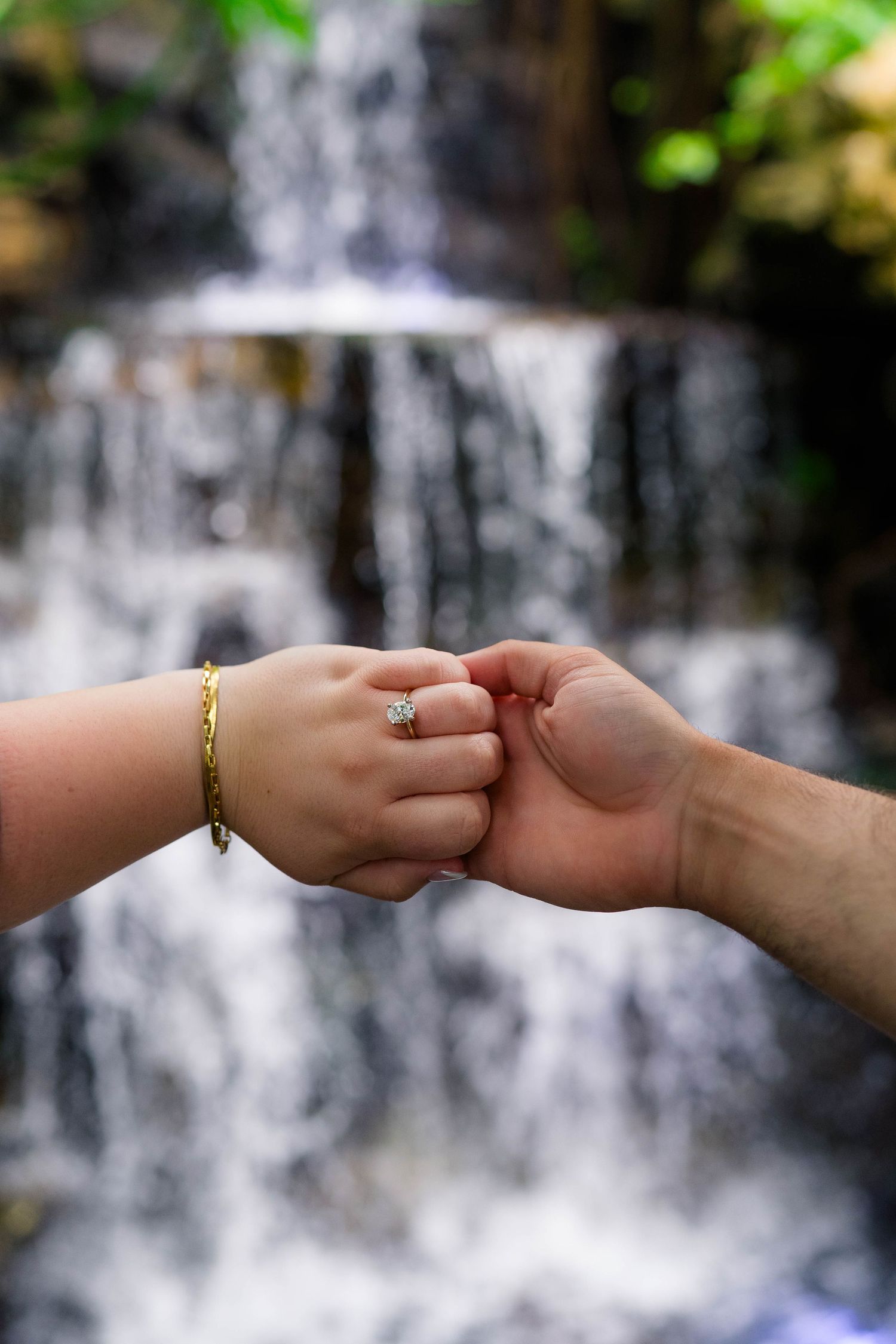 Couple holding hands with engagement ring and gold bracelet in front of a blurred waterfall backdrop.