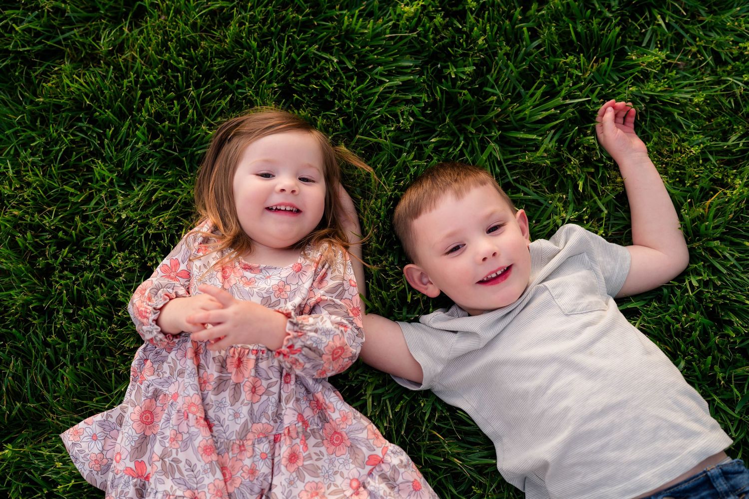 Two young children, a girl in a floral dress and a boy in a gray shirt, smiling while lying on green grass.