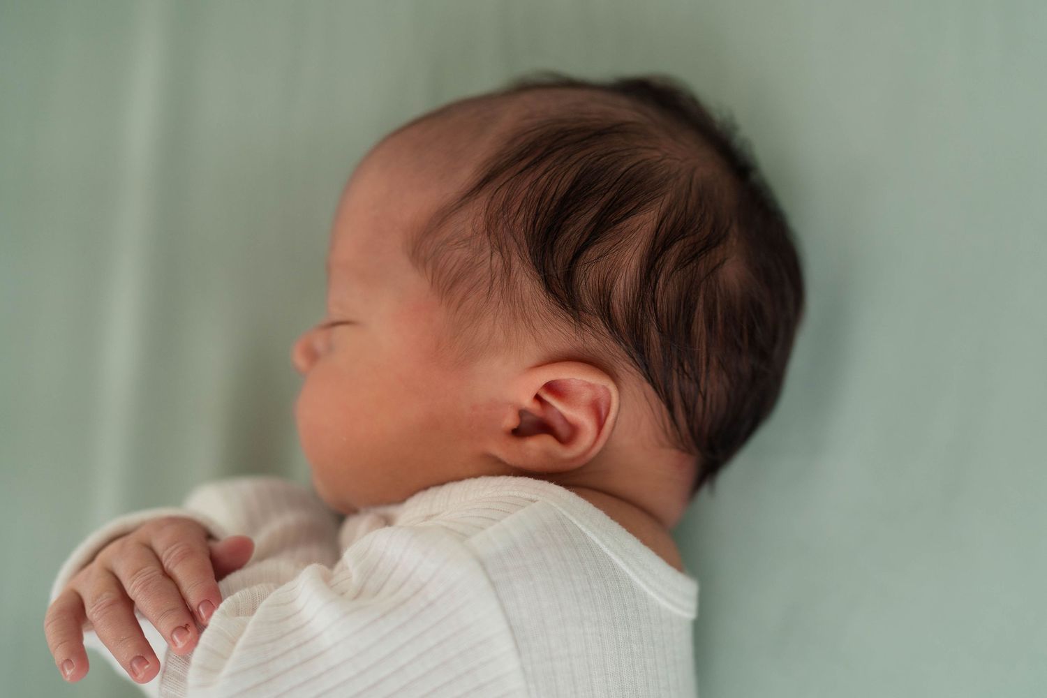 Close-up of a newborn baby with dark hair sleeping peacefully in a white outfit.