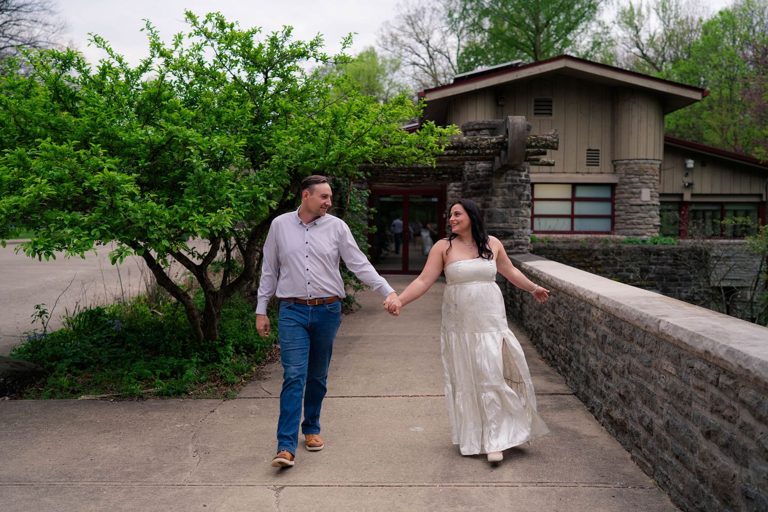 Engaged couple holding hands and walking across a stone bridge with a rustic building and green trees behind them.